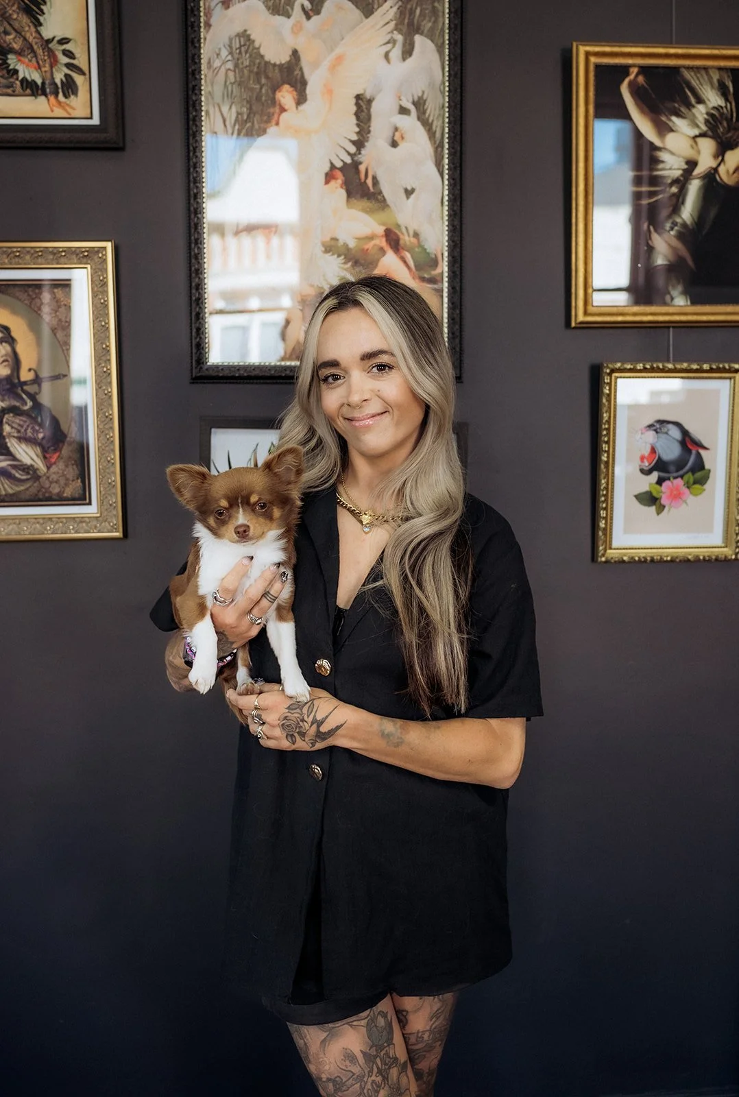 Smiling tattooed woman in a black shirt gently holding a small brown and white dog against a gallery wall of framed artwork