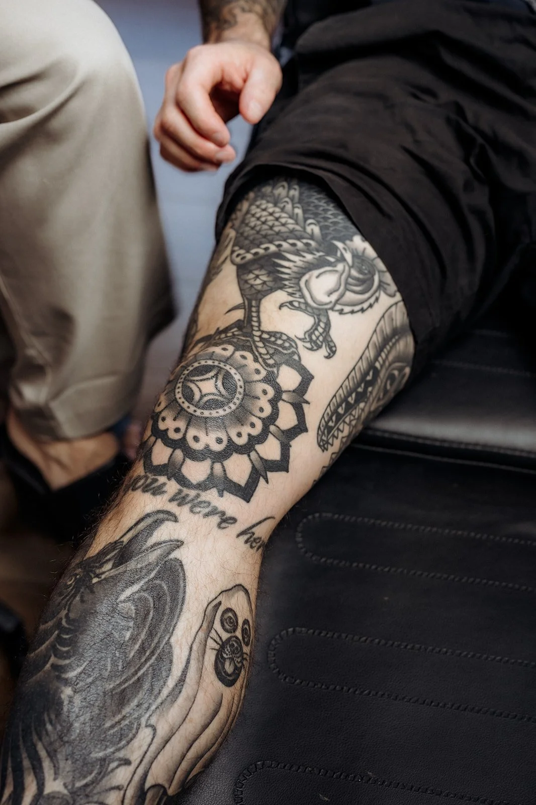 Close-up of a tattooed forearm resting on a treatment chair, featuring detailed black ink designs including a mandala, bird, and script lettering