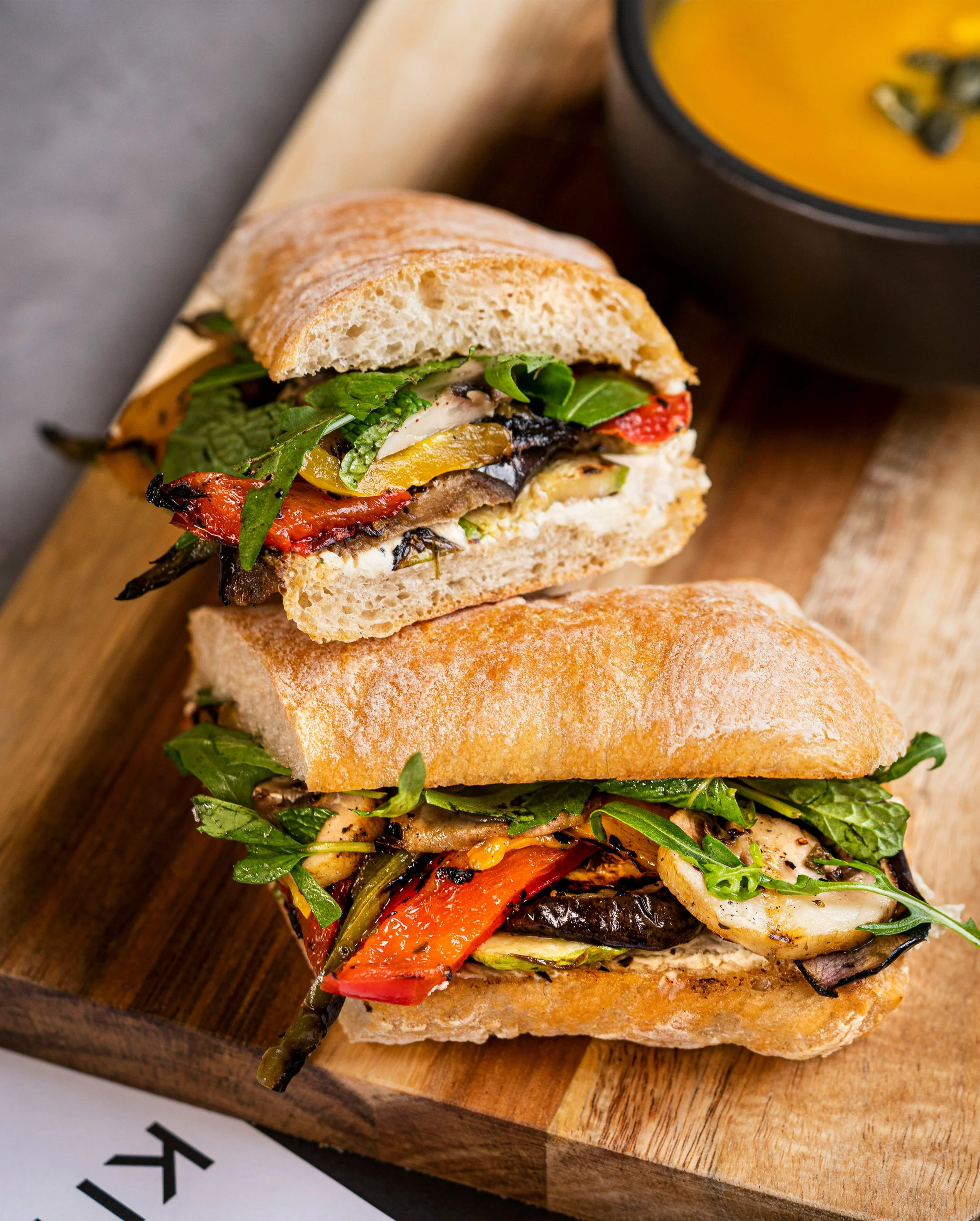 A sandwich with roasted vegetables and greens on a wooden tray, with a bowl of soup in the background.