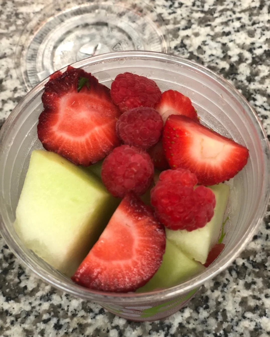 A plastic cup filled with sliced strawberries, raspberries, and chunks of green apple on a speckled gray countertop.