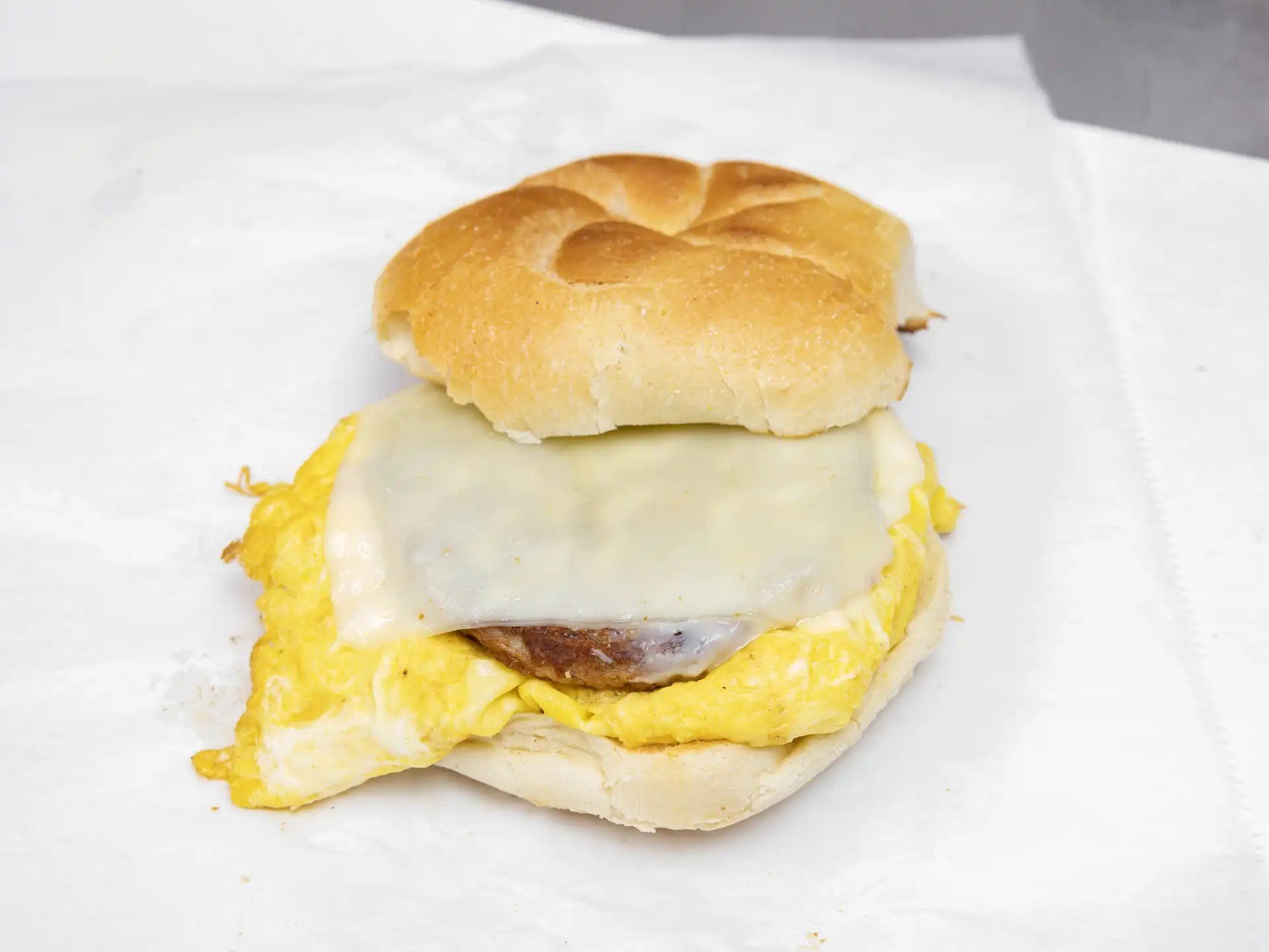 Breakfast sandwich with scrambled eggs, melted cheese, sausage patty, and a toasted bun, placed on white parchment paper.