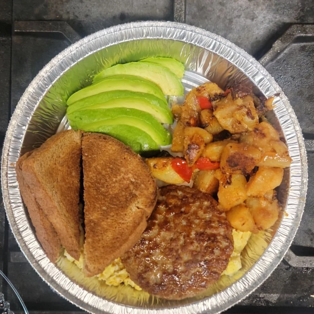 Aluminum tray filled with sliced avocado, breakfast sandwich with toasted bread, cooked breakfast sausage patty, scrambled eggs, and roasted diced potatoes with peppers.