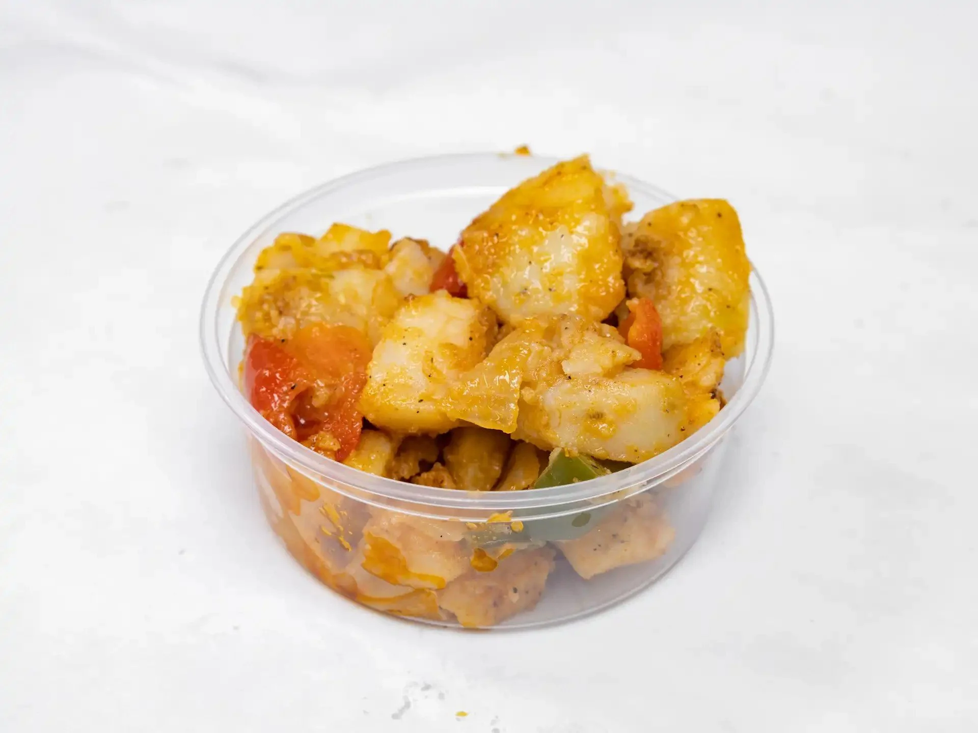 A small clear plastic cup filled with fried potato chunks and peppers on a white surface.
