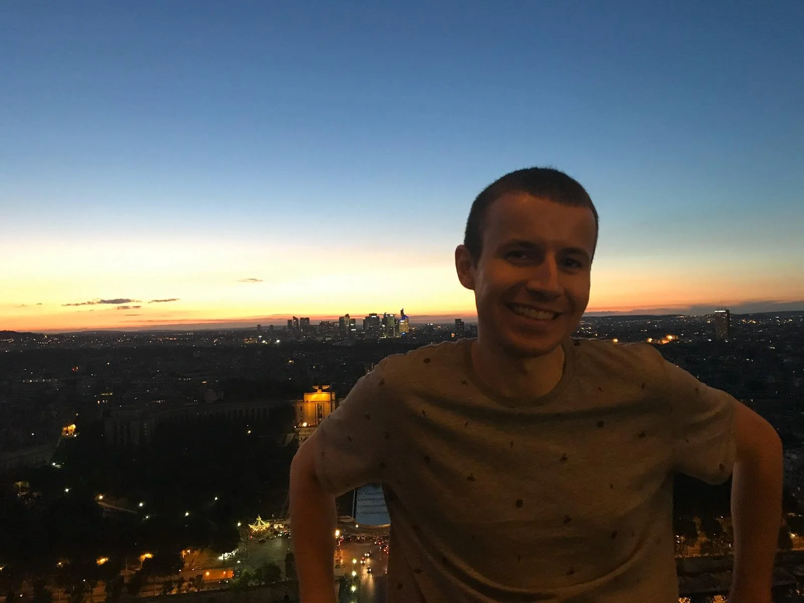 A young man smiling at sunset on a rooftop overlooking a city skyline, with a clear sky transitioning from blue to orange.