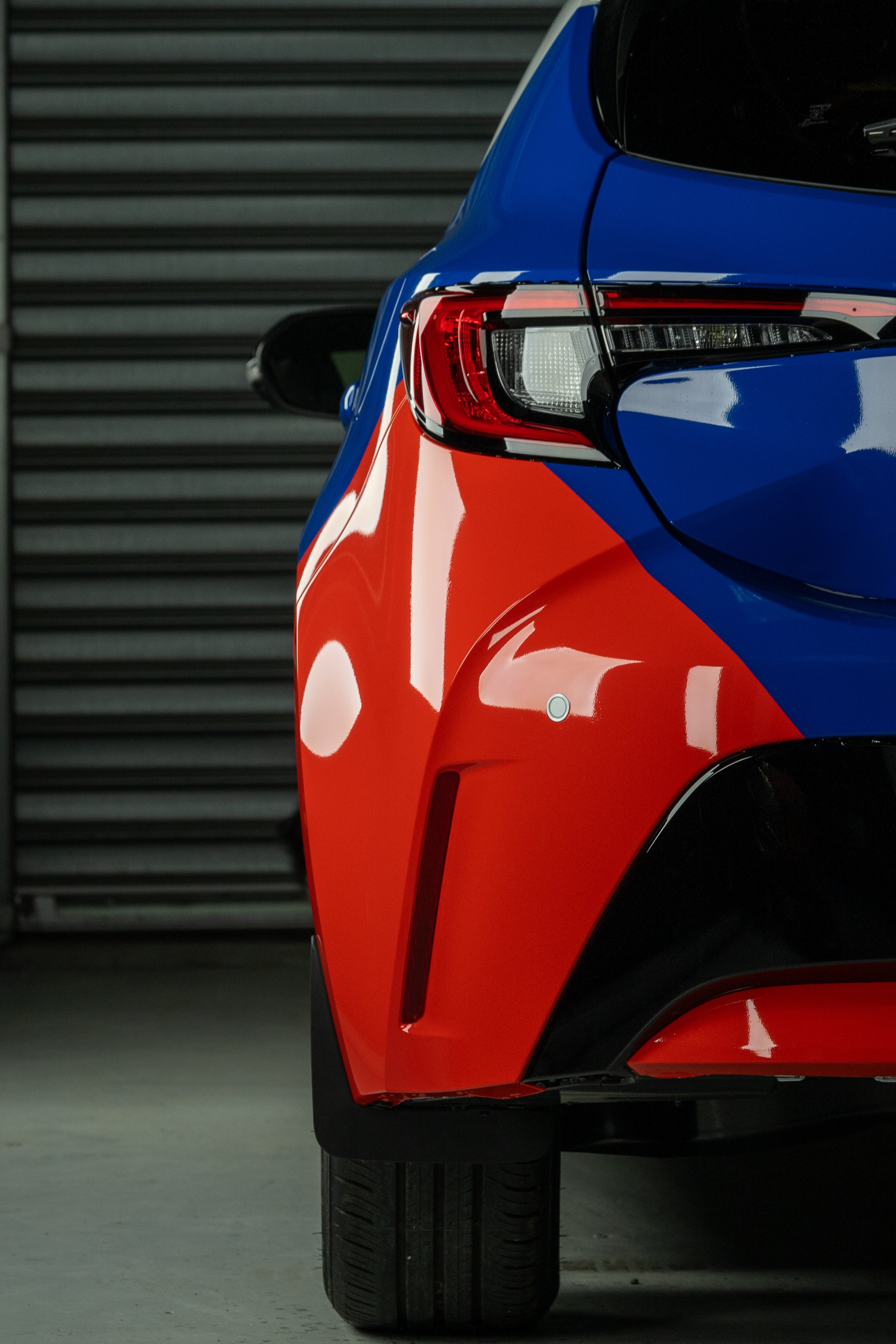 Close-up of the rear corner of a red and blue sports car, showing tail light, rear wheel, and part of the side panel, with a gray garage door in the background.
