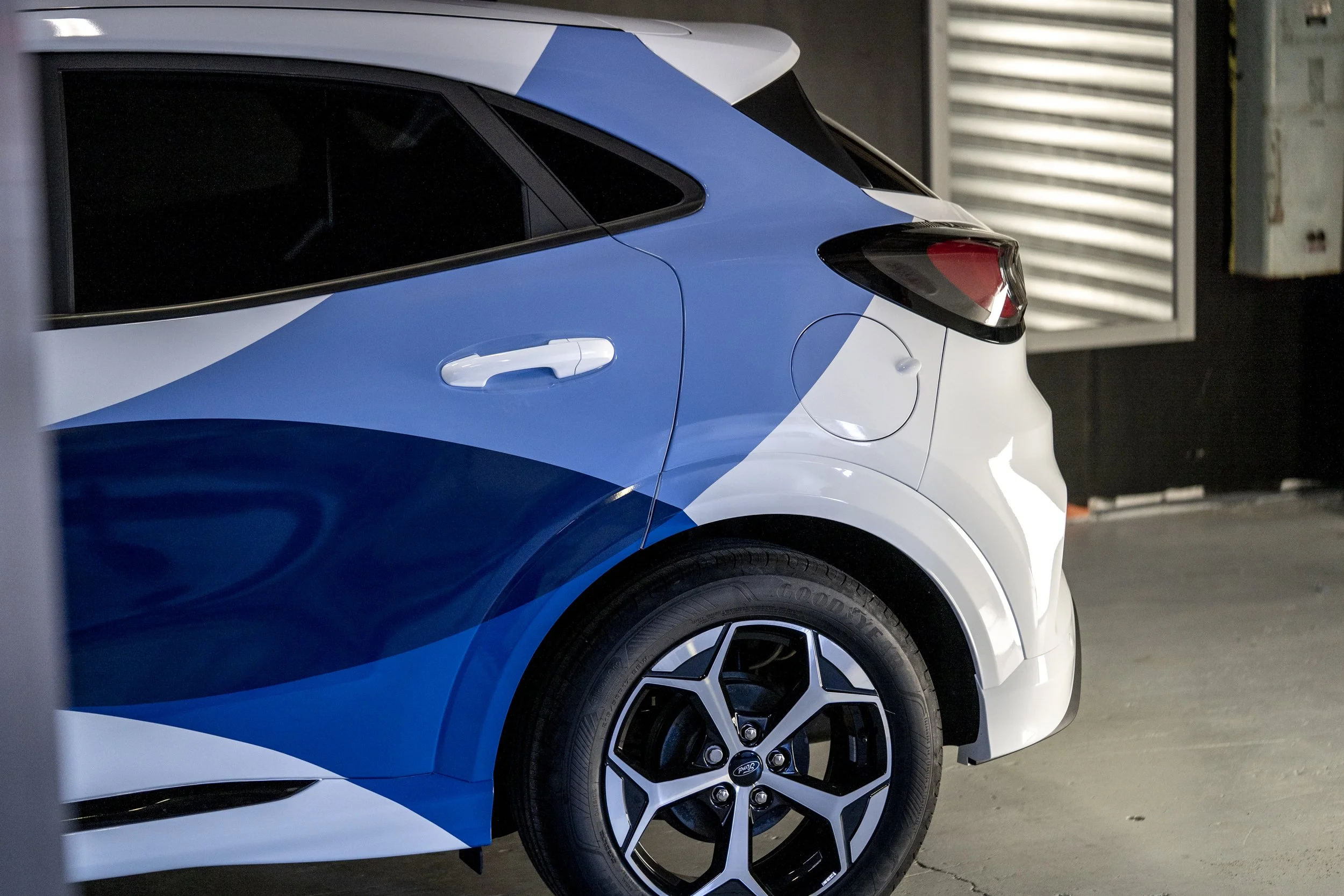 Close-up of a modern electric vehicle, showing part of the front and side with a blue and white color scheme, black wheels, and a parking garage background.