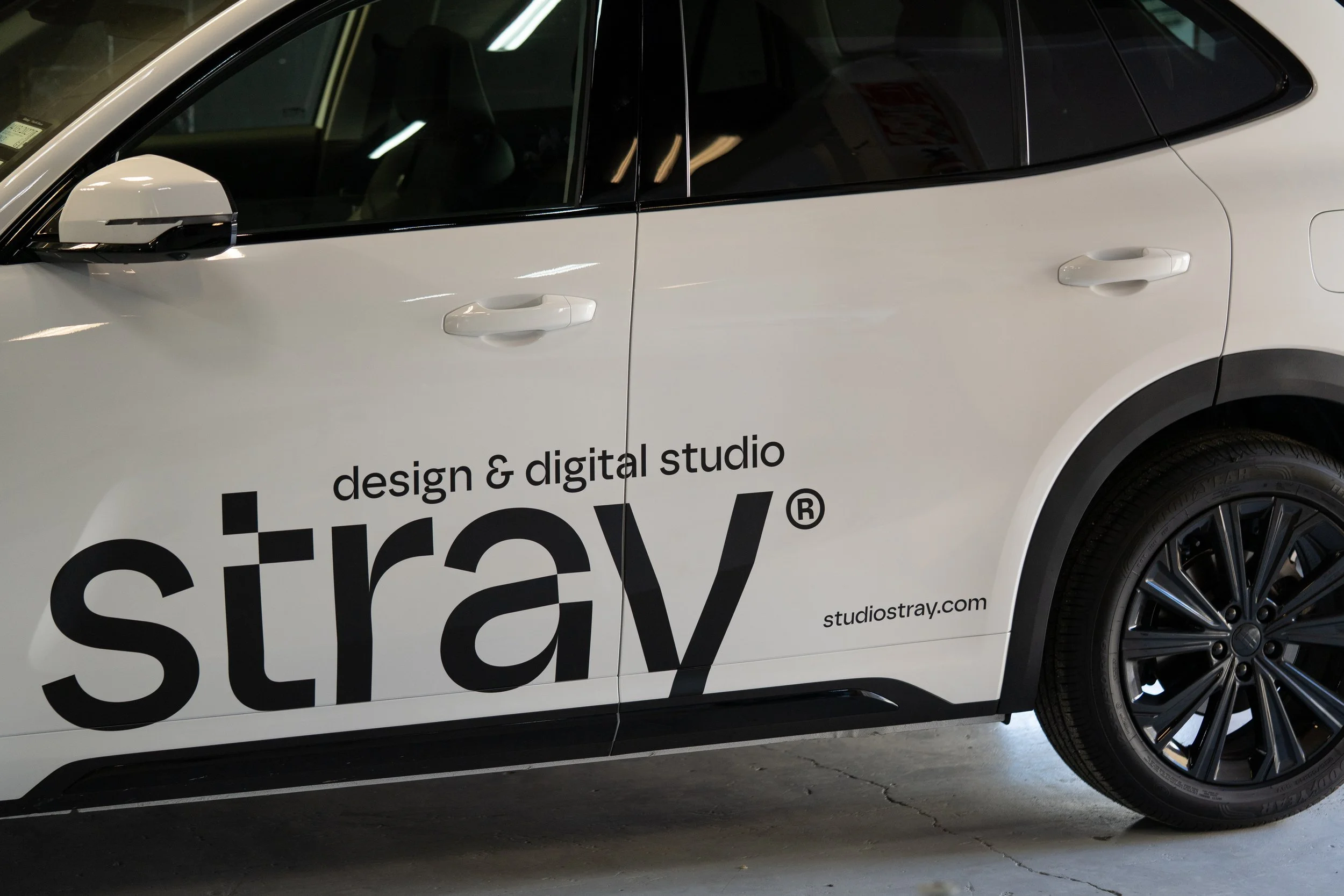 Partial view of a white SUV with black tinted windows and black wheels, featuring a large black and white logo and text for 'studio stray' on the side.