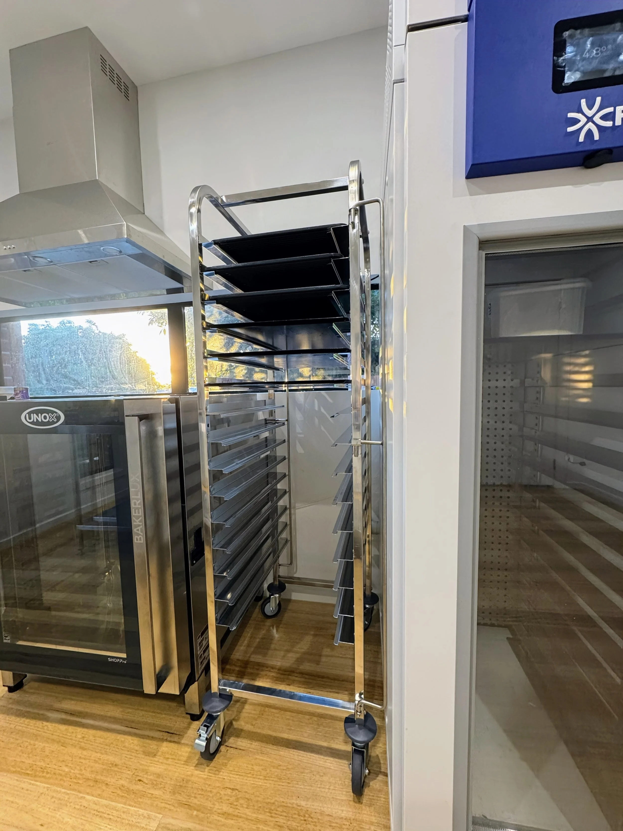 Empty bakery cooling rack cart with multiple black trays, standing on a wooden floor in a commercial kitchen.