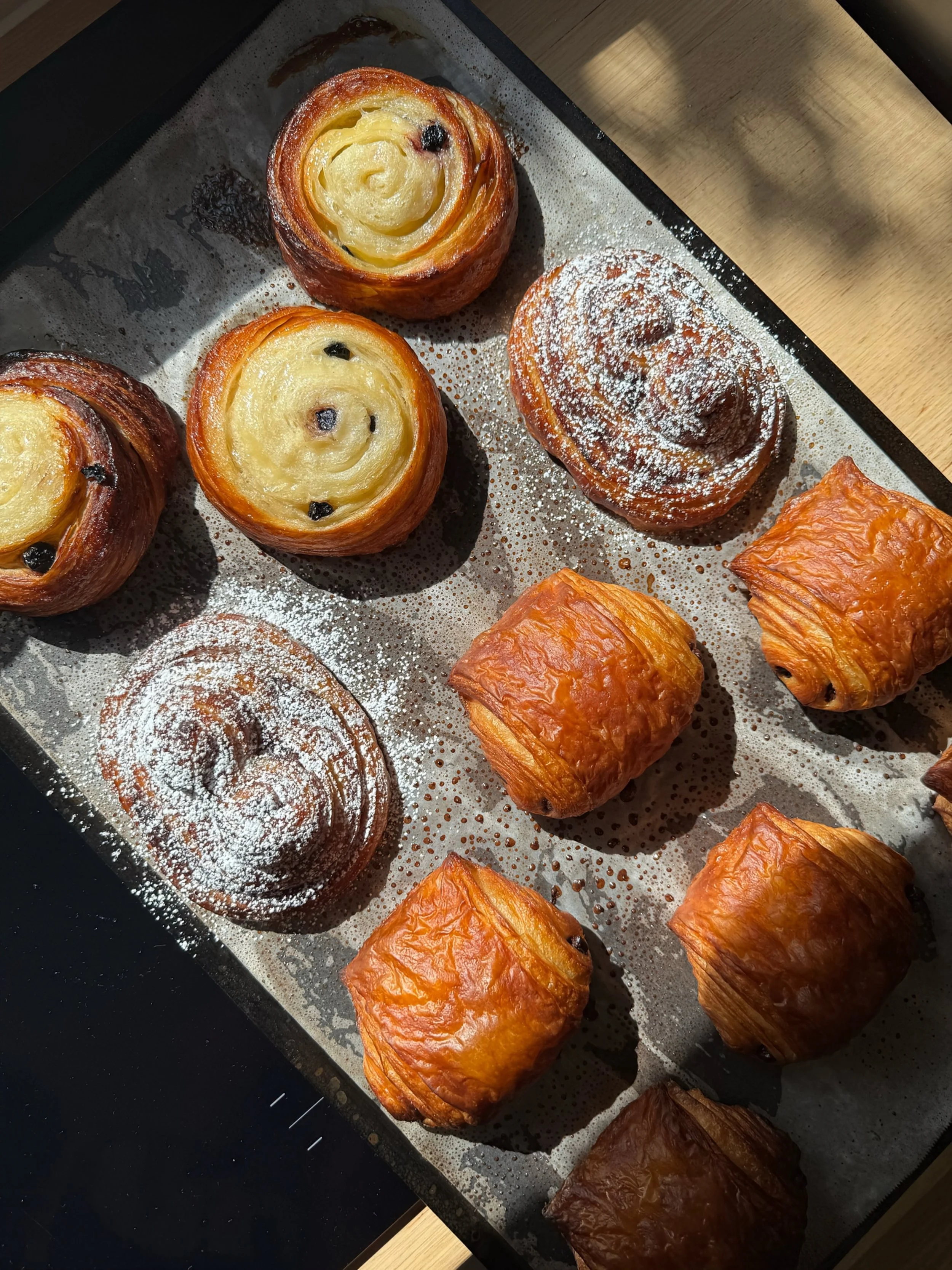 Assorted freshly baked pastries on a baking sheet, including Danish pastries with swirled tops, chocolate chips, dusted cinnamon rolls, and puff pastry squares.