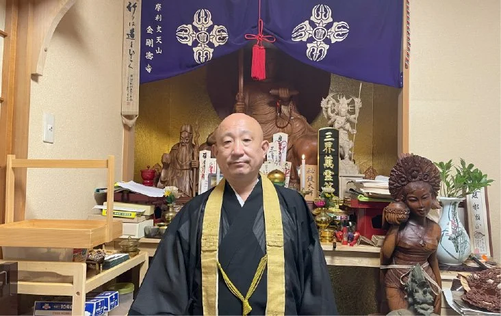仏教僧侶が仏壇の前で写真を撮っている。背景には仏像とお守り、供物が置かれている。
