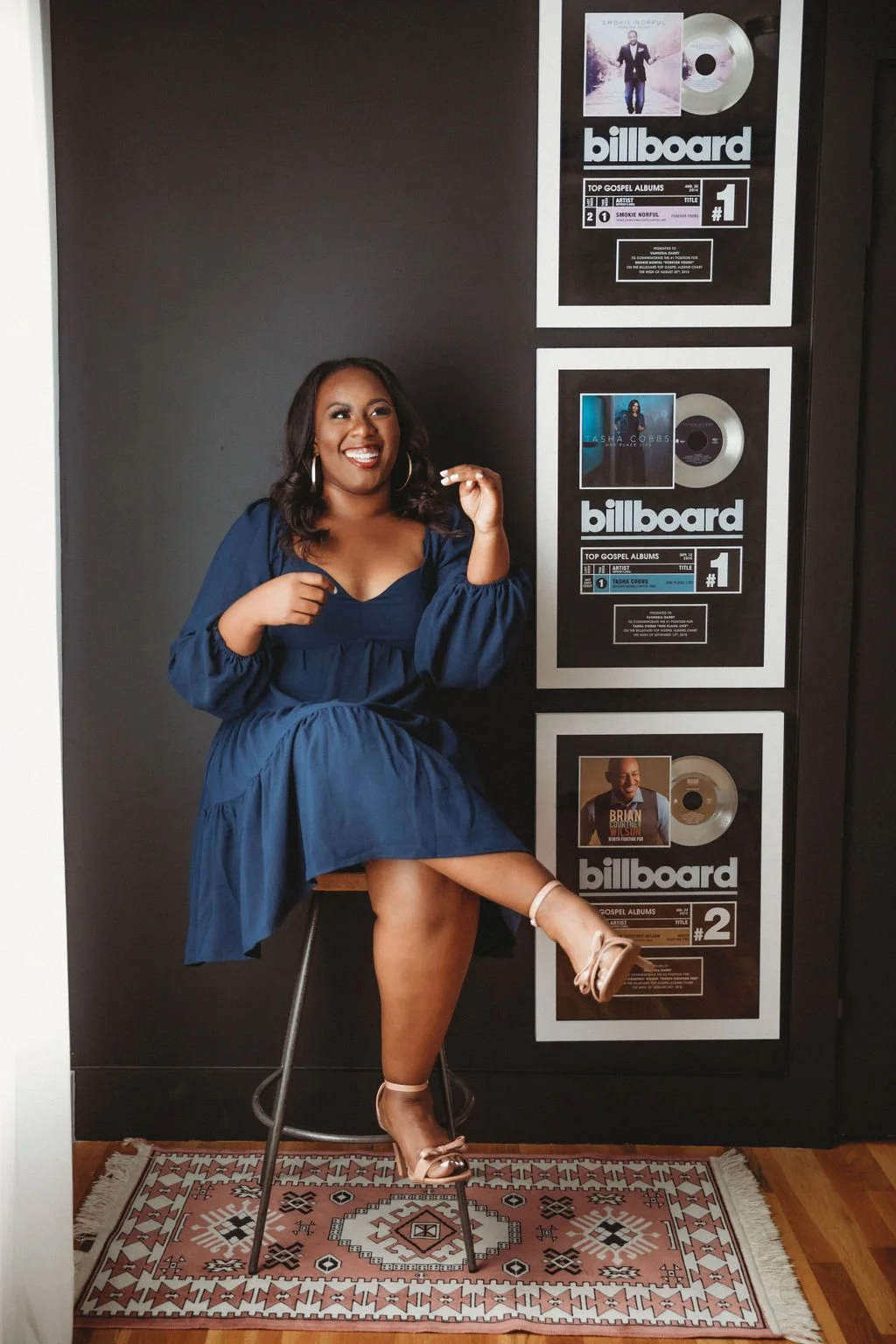 A woman in a blue dress sits on a stool, smiling and posing with one leg raised, in front of a black wall decorated with framed Billboard award plaques for top gospel albums.