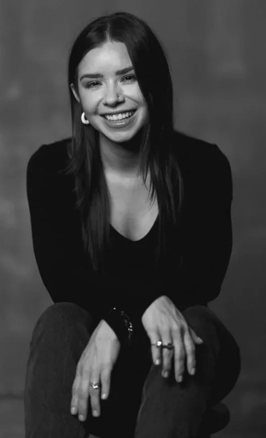 A black and white portrait of a young woman with long dark hair, smiling, wearing a dark top and sitting with legs crossed.