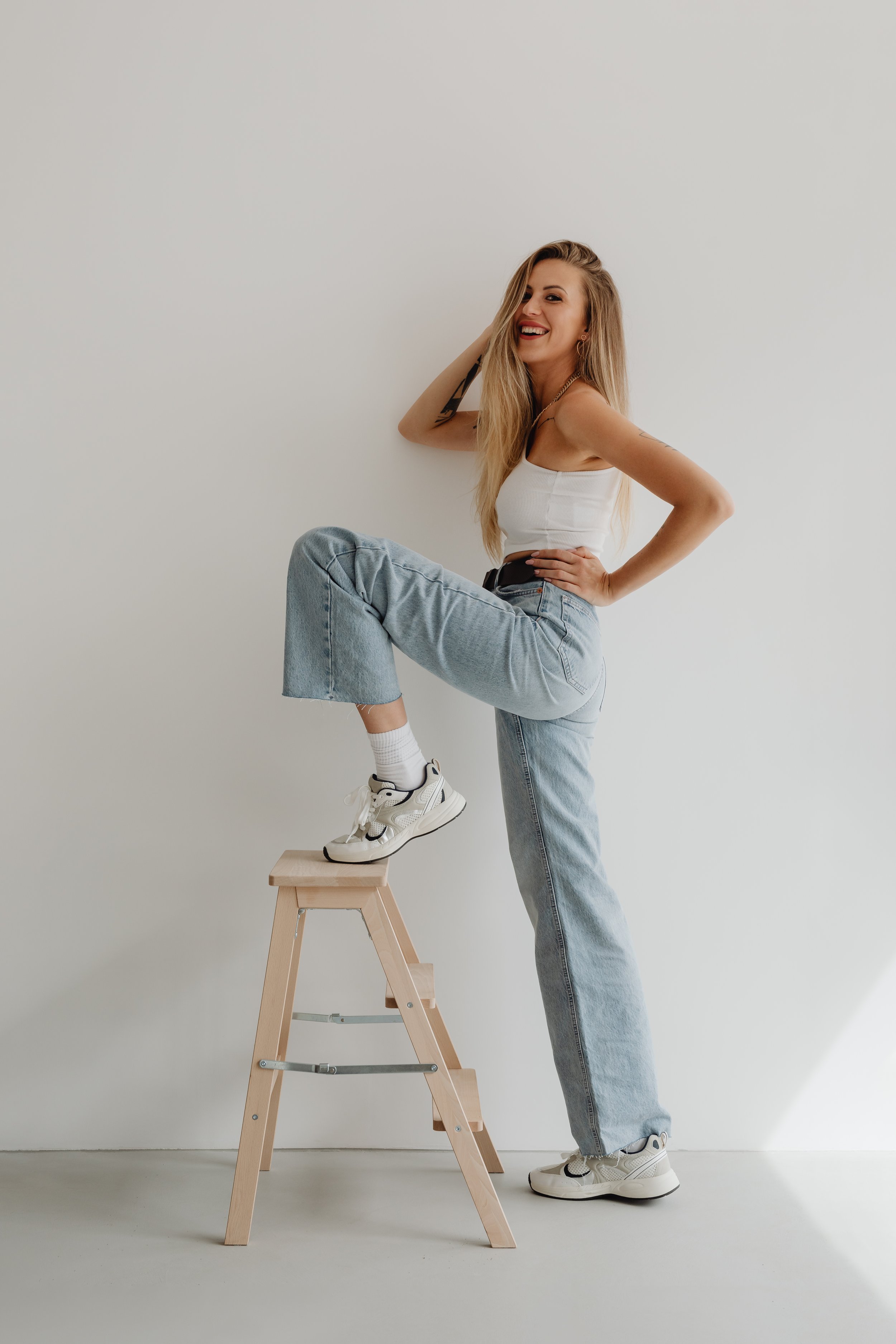 A young woman with long blonde hair, wearing a white tank top, light blue jeans, and sneakers, poses next to a wooden stool. She has a big smile, with one hand on her hip and the other touching her head.
