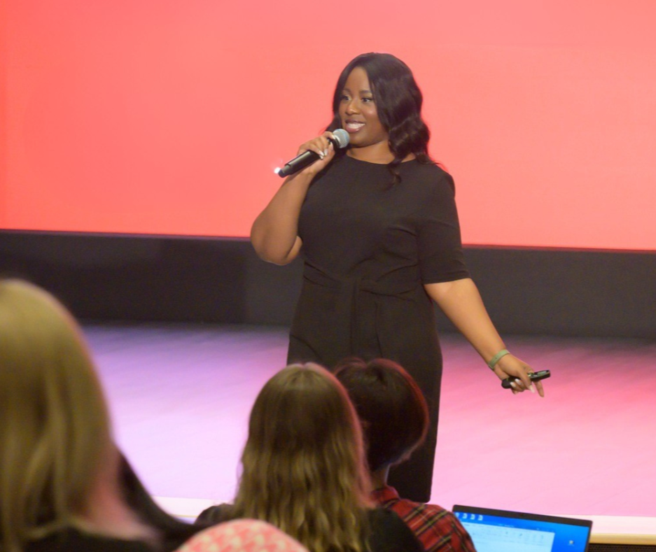 A woman in black clothing standing on stage, holding a microphone and speaking to an audience.