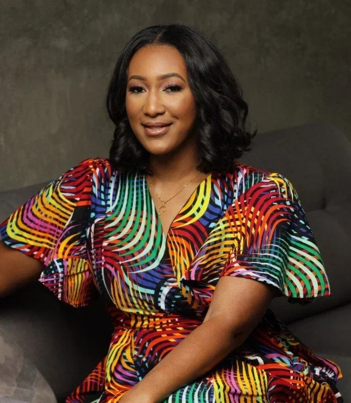 A woman with black wavy hair wearing a colorful, patterned dress, sitting on a dark couch against a muted background.