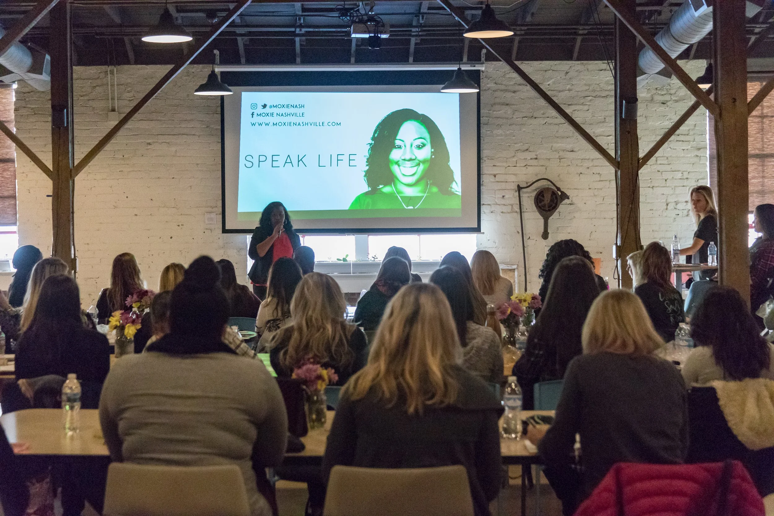A woman speaking at a conference or seminar in a large industrial-style room with exposed brick walls, wooden beams, and a projection screen displaying a smiling woman's portrait with the text 'Speak Life'. The audience is seated at round tables with flower centerpieces, and some people are drinking bottled water.