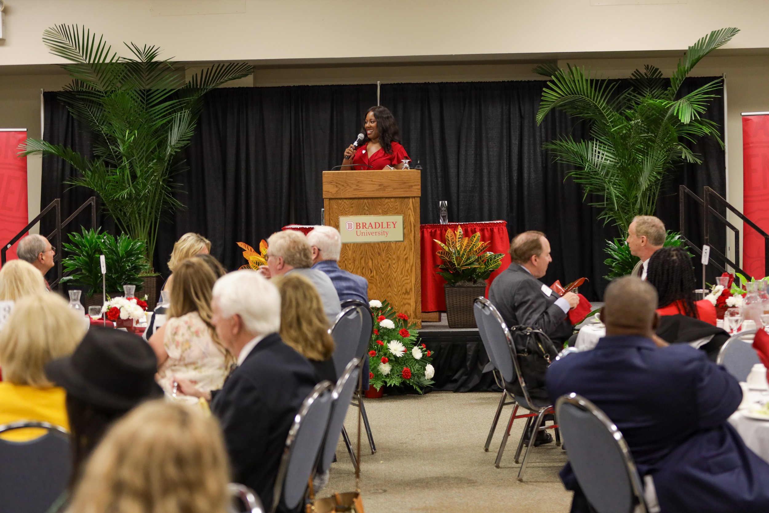 A woman in a red dress stands at a podium speaking into a microphone at an event at Bradley University. The stage is decorated with large green plants and flowers, and there are attendees seated at round tables in the foreground.
