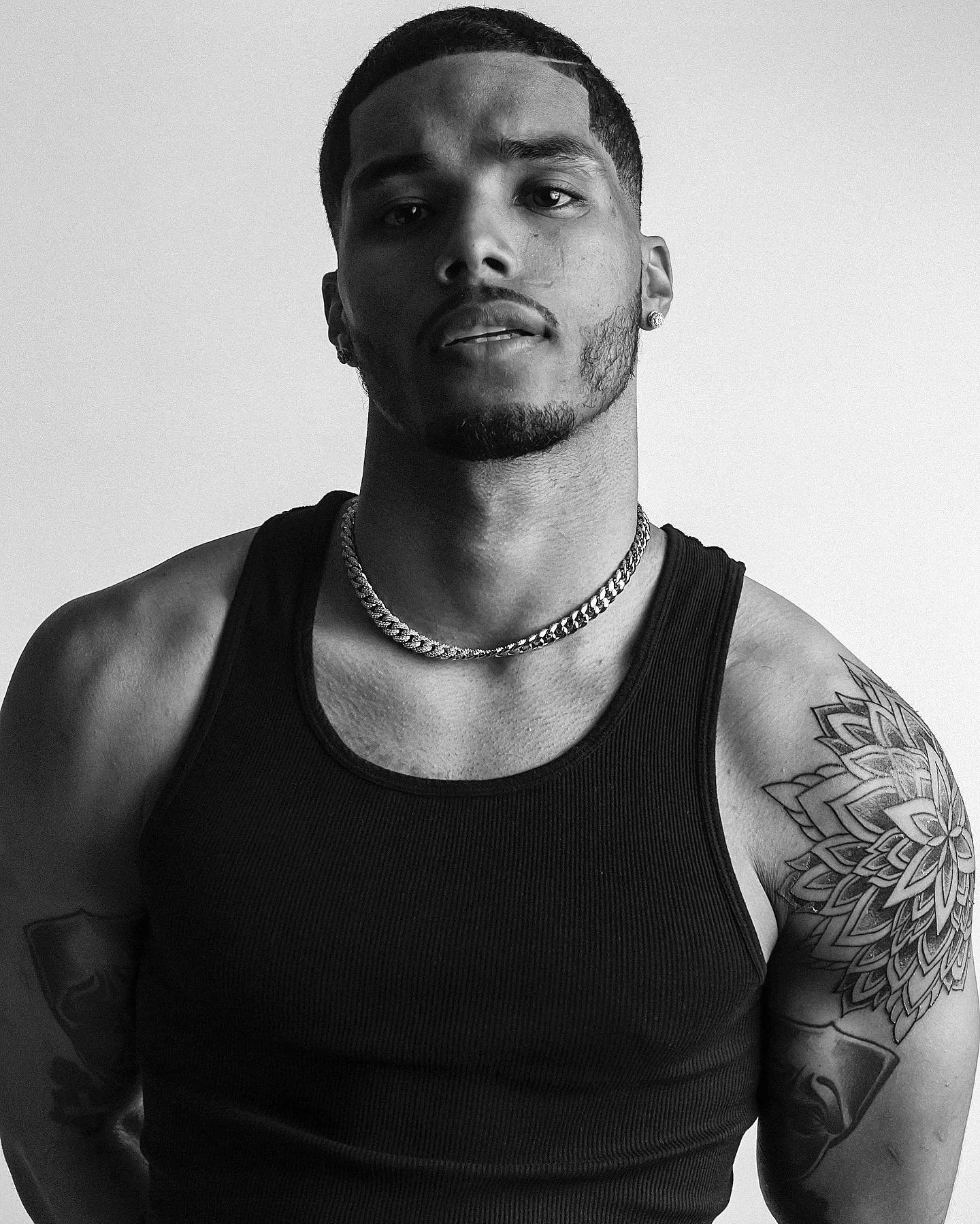 Black and white photo of a young man with short dark hair and facial hair, wearing a sleeveless black shirt, two necklaces, and earrings. He has tattoos on his left arm and right shoulder. The background is plain and light-colored.