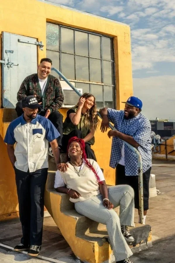Group of five friends on a rooftop, smiling and joking, with a bright yellow building and cloudy sky in the background.