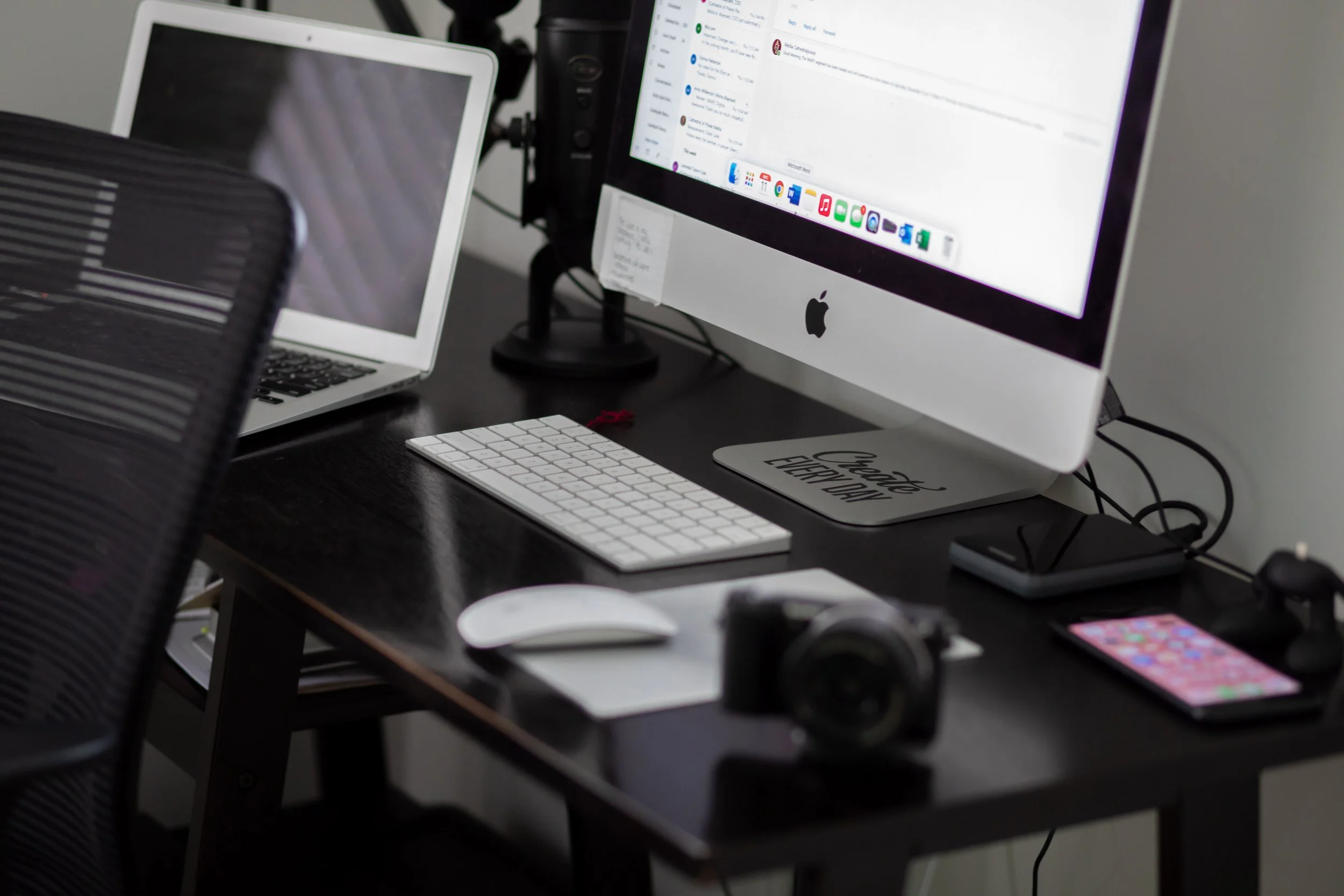 An office desk setup with an Apple iMac monitor displaying an email or messaging app, a laptop to the left with a reflective screen, a white keyboard, a mouse, a smartphone, and various electronic accessories on the dark desk surface.