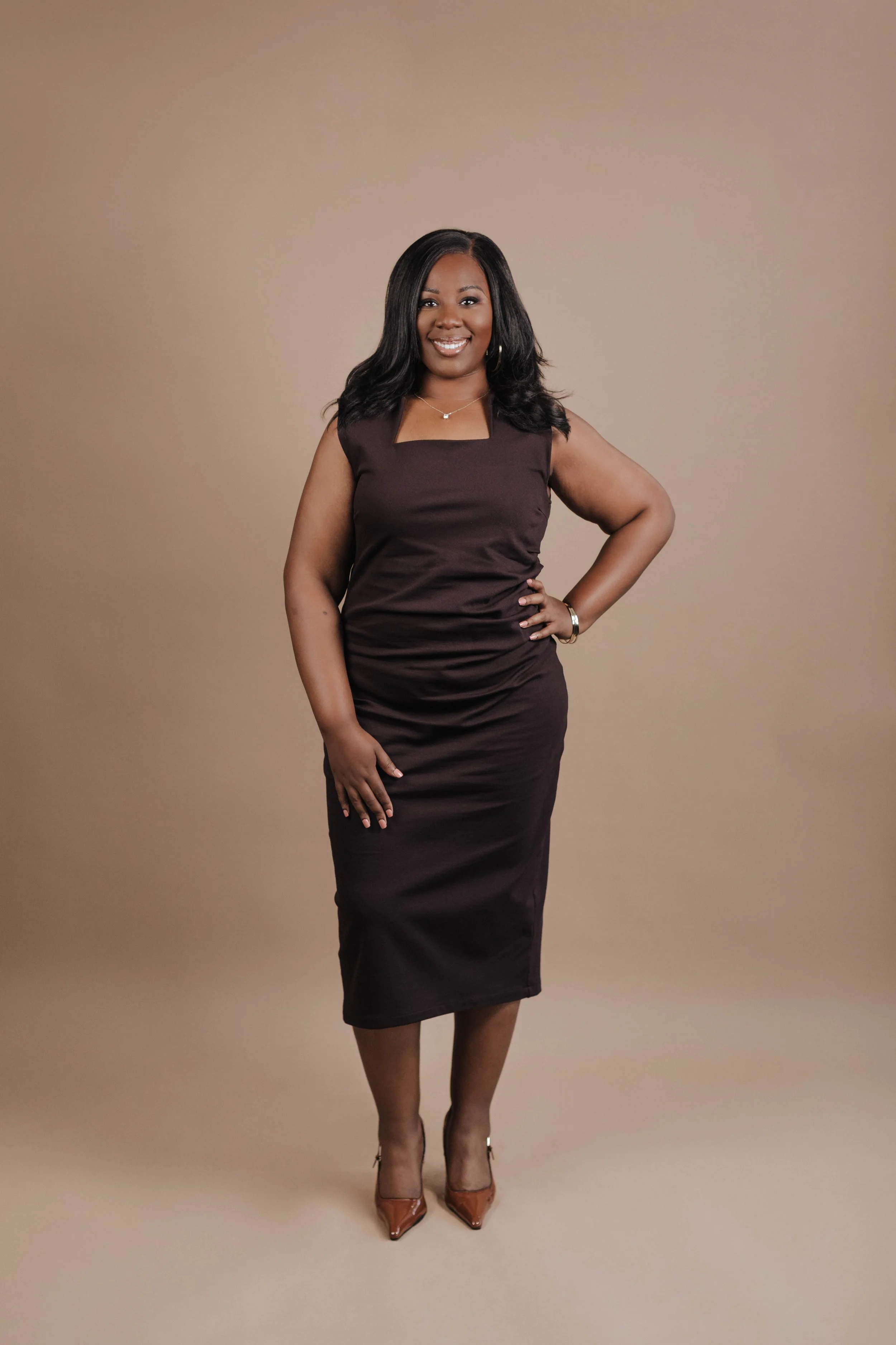 A woman in a fitted dark brown dress with a square neckline, wearing high heels, standing against a beige background, smiling confidently with one hand on her hip.