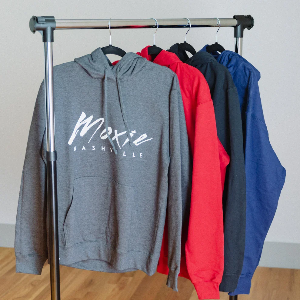 Four hooded sweatshirts hanging on a metal clothing rack inside a room with hardwood floors. The gray sweatshirt in the front has the words 'Moxie Nashville' in white text. Behind it are red, black, and blue hoodies.
