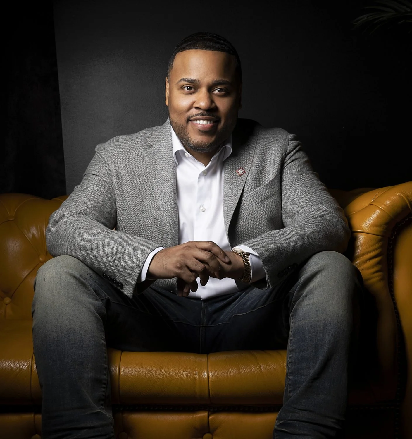 A man with short hair and a beard, dressed in a gray blazer and white shirt, seated on a mustard-colored leather sofa against a dark background.