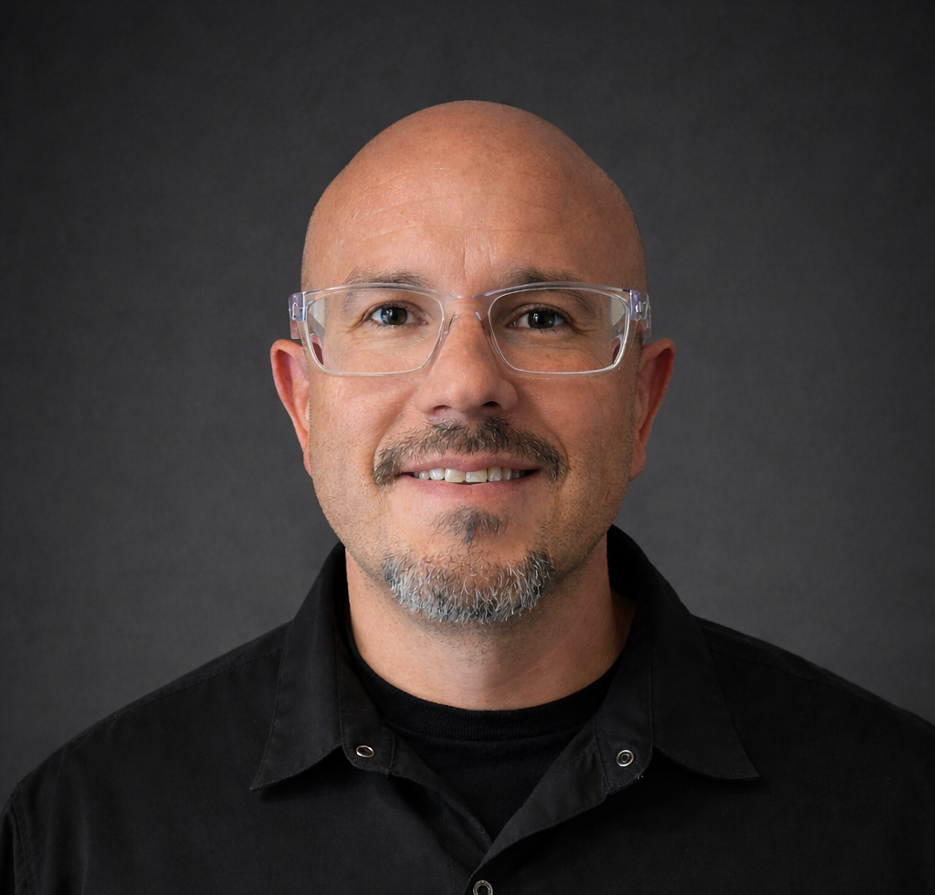 Close-up portrait of a bald man with glasses, a goatee, wearing a black shirt, smiling against a dark background.
