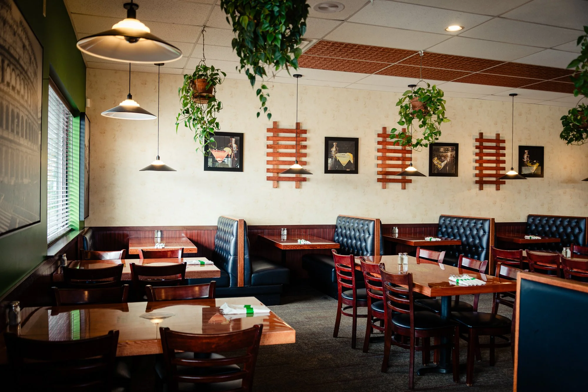 Empty restaurant with wooden tables, dark blue cushioned booths, brown chairs, hanging lights, and green plants.