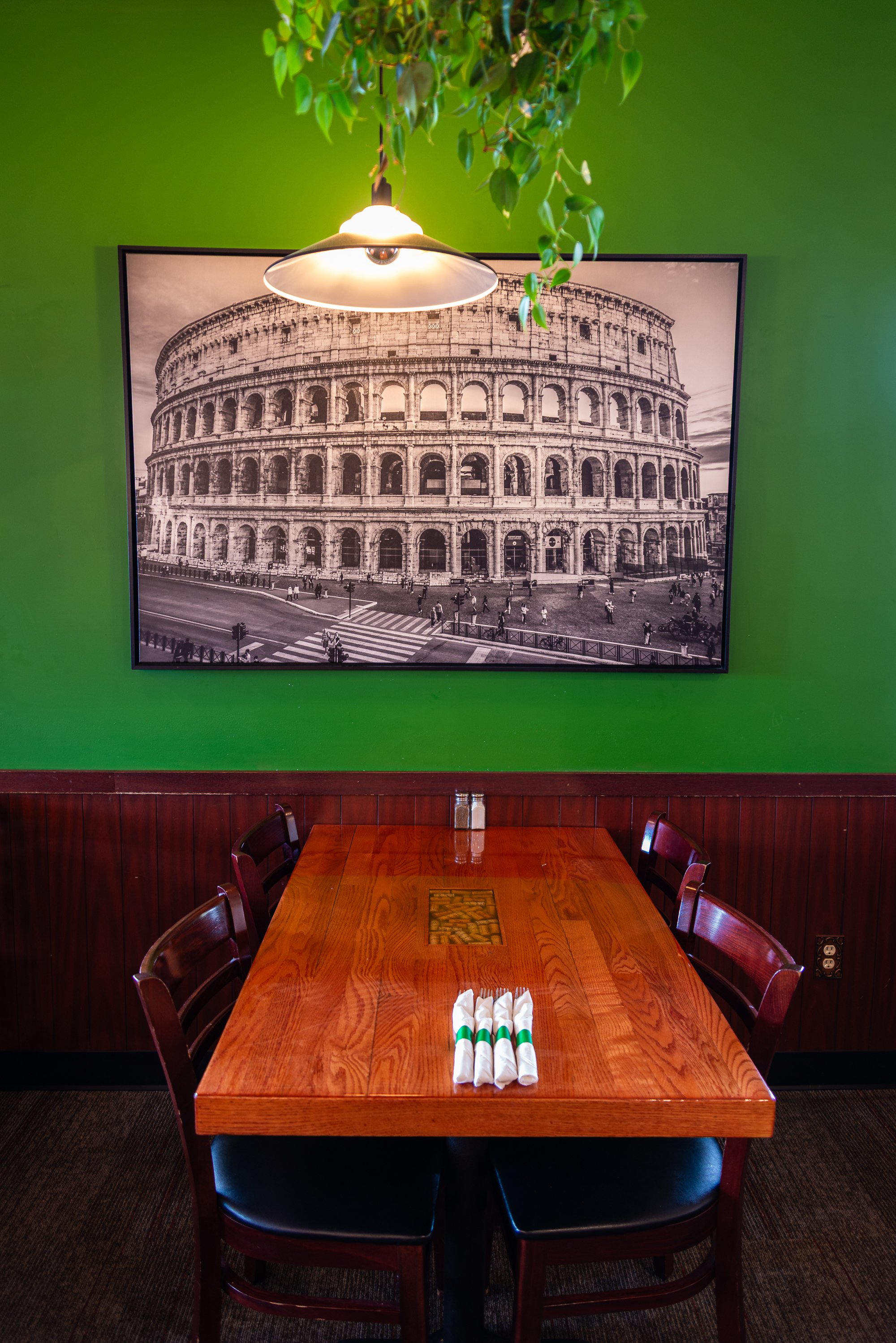A wooden dining table set for four with folded white napkins and utensils, positioned in front of a green wall with a large black-and-white photo of the Roman Colosseum, a hanging light fixture, and a plant above.