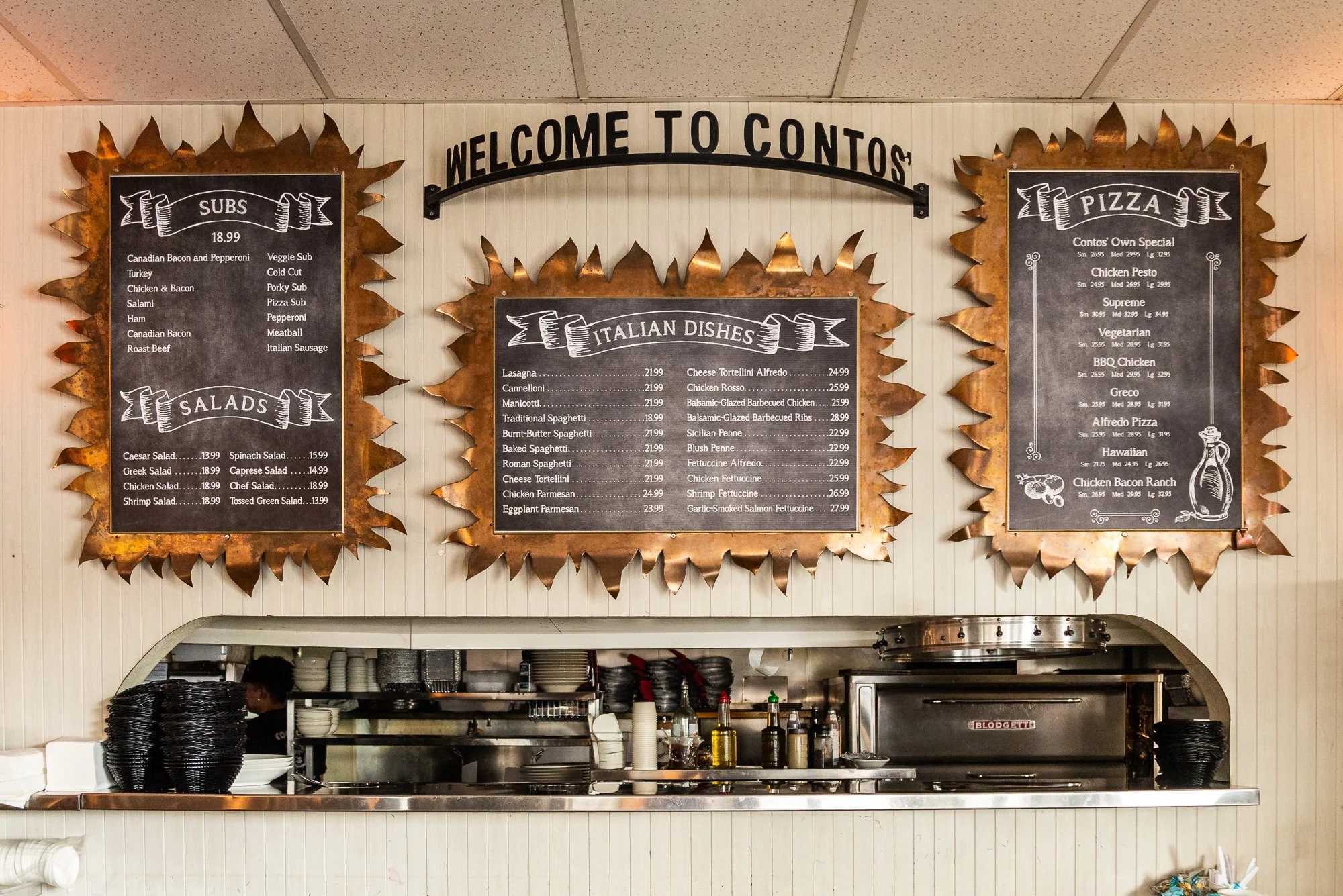 Restaurant menu boards with a copper leaf border, displaying Italian dishes, salads, and pizza options, with a kitchen window below.