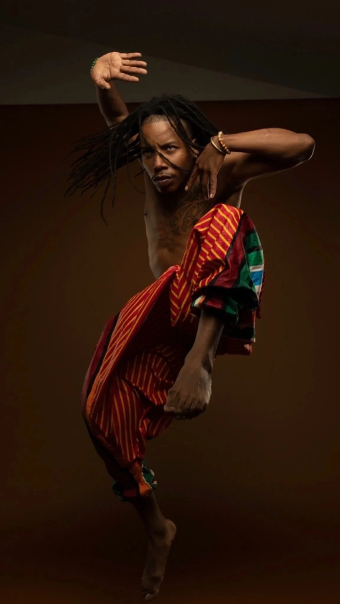 Person with long dreadlocks, wearing colorful striped pants, performing a dance pose against a dark background.