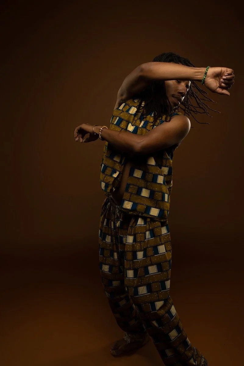 A person with dreadlocks wearing patterned traditional African clothing, striking a dance pose with arms crossed and one arm covering part of their face, against a brown background.