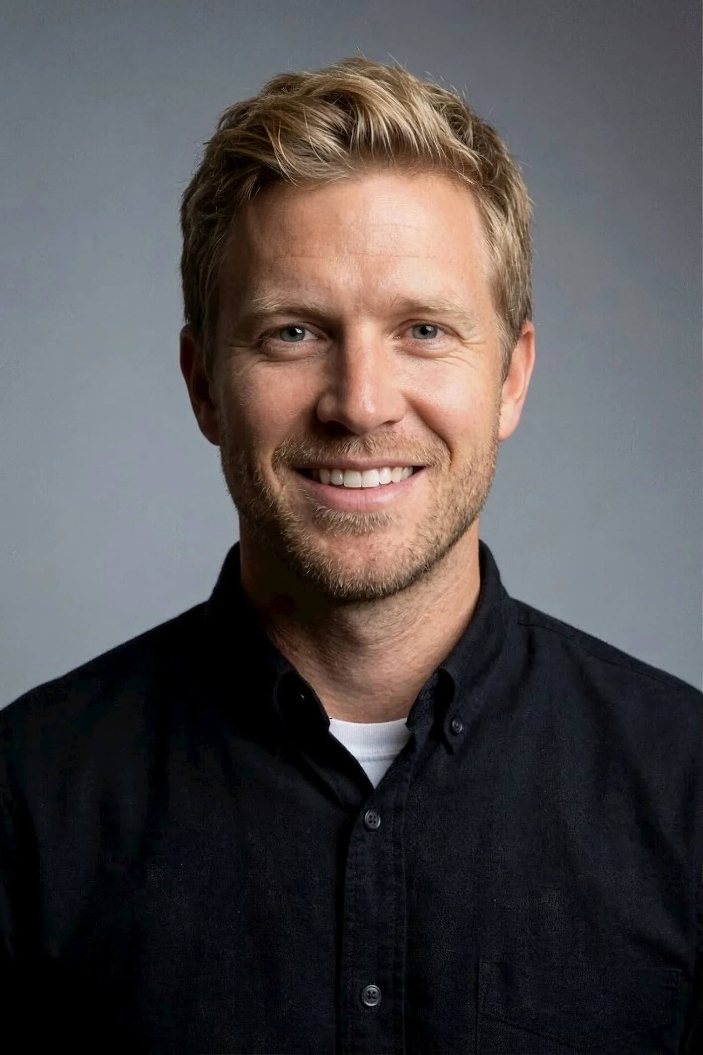 Portrait of a smiling man with blonde hair, wearing a black collared shirt and a white undershirt, against a gray background.