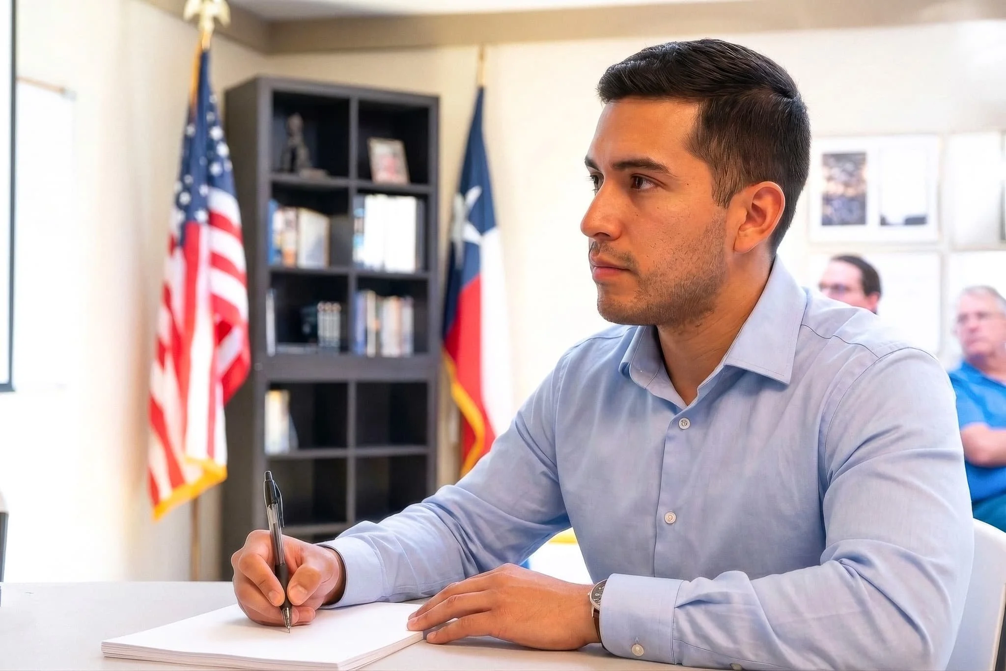 A man with dark hair, wearing a light blue shirt, is sitting at a desk, writing on a notepad with a pen. Behind him, there are two flags, one American and one possibly Texas, and a bookshelf with decorative items and framed pictures. There are also o