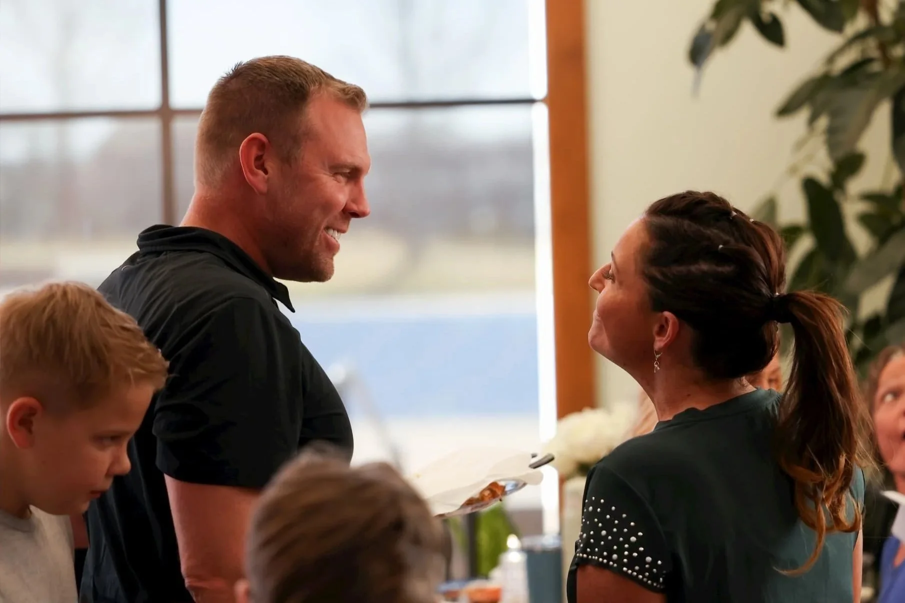 A man with short reddish hair and a beard is smiling while looking at a woman with long dark hair tied in a ponytail, who is looking up at him. They appear to be in a cheerful, indoor setting, possibly a social or celebratory event.
