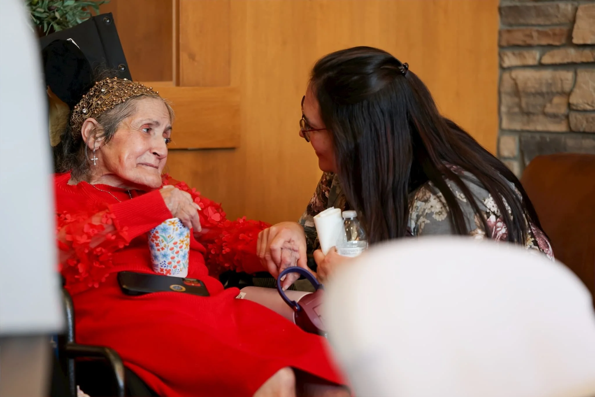 An elderly woman with long gray hair, wearing a golden beaded headband, is sitting in a wheelchair. She is holding hands with a woman with long dark hair, glasses, and a floral top. They are engaging in a heartfelt moment in a room with wood-paneled 