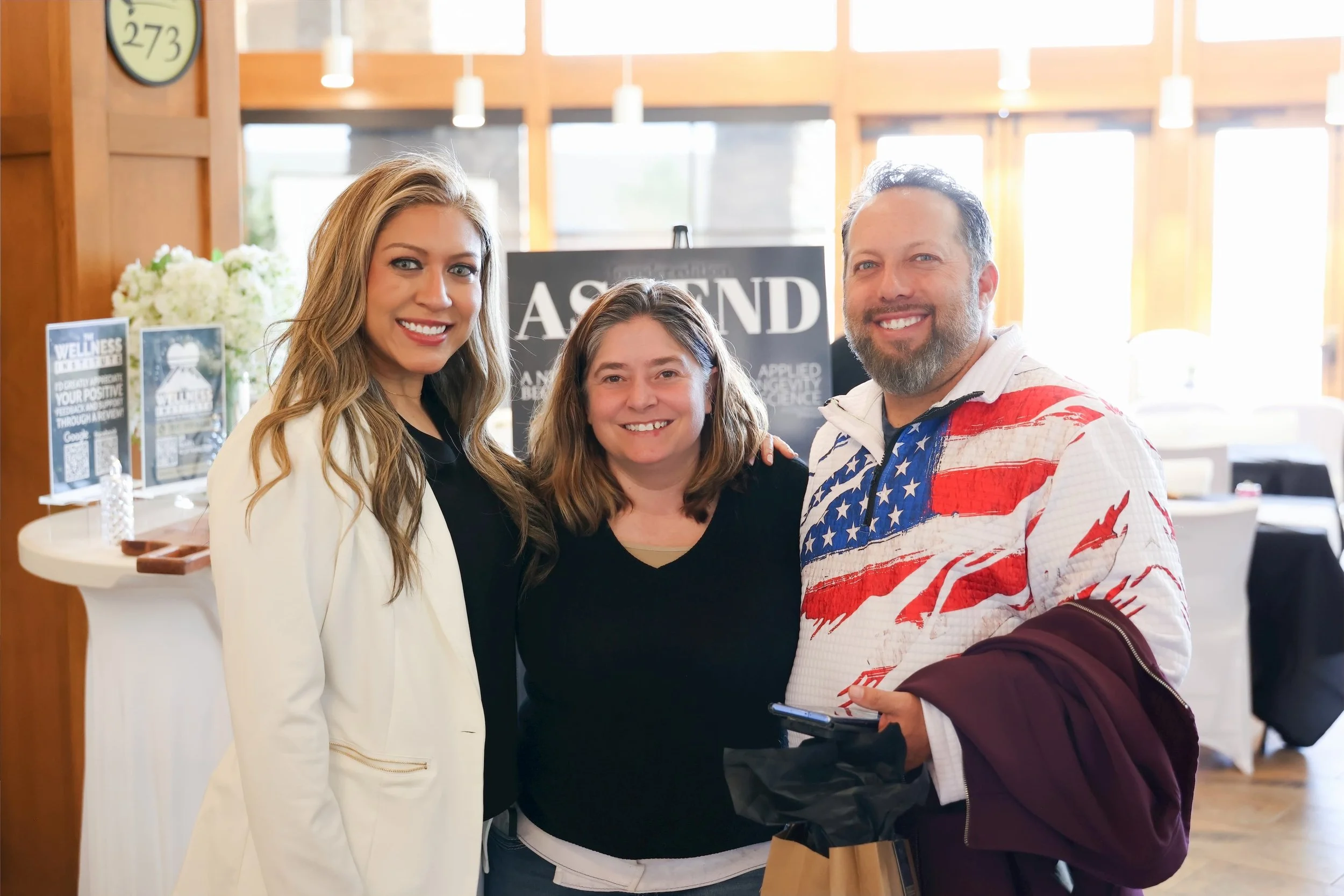 Three smiling people posing indoors at a social event, with a woman in a white blazer on the left, a woman in a black shirt in the middle, and a man in a patriotic shirt on the right.