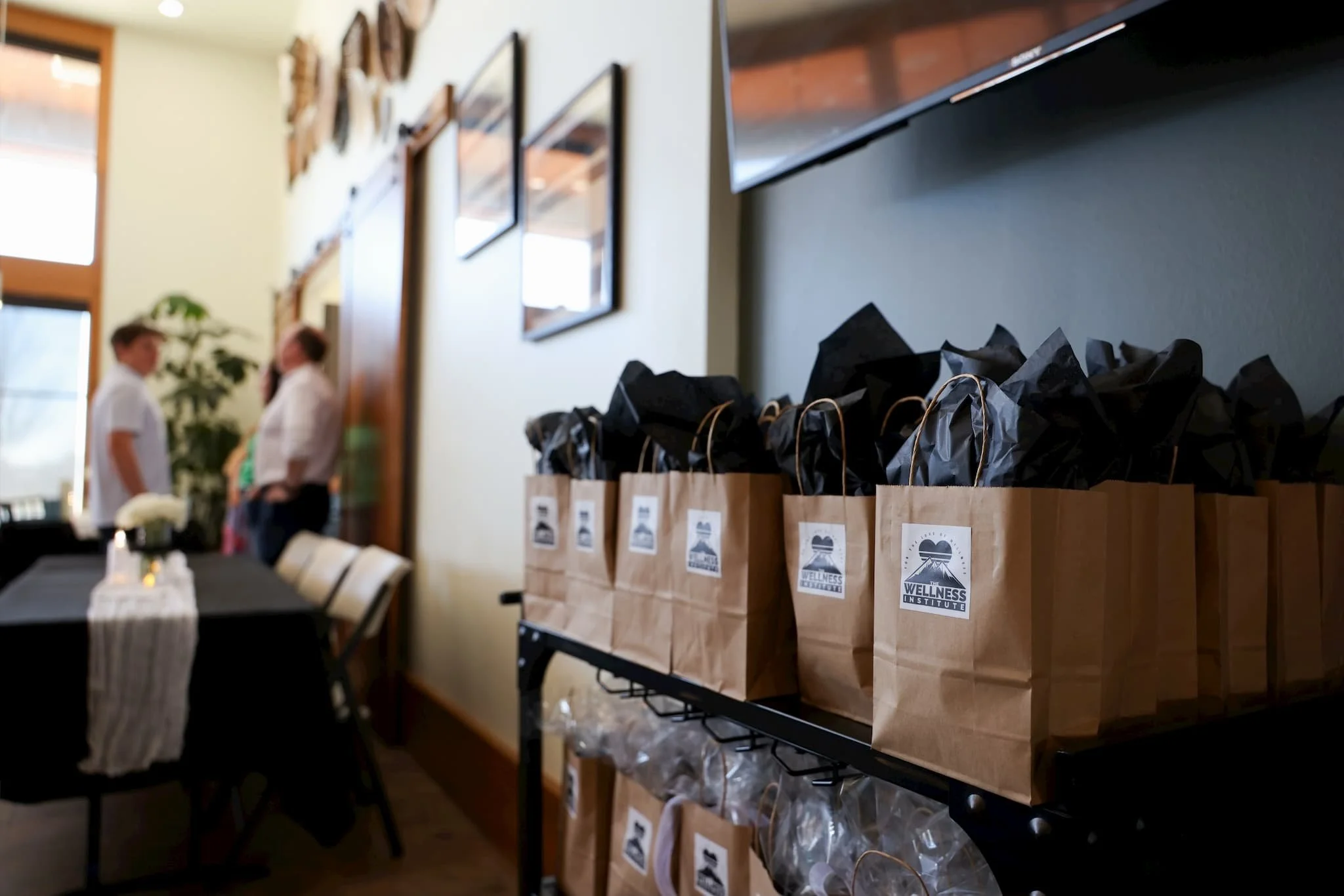 Brown paper bags with black tissue paper inside on a shelf, with a logo reading 'The Wellness Institute' and an image of mountains and a sun.