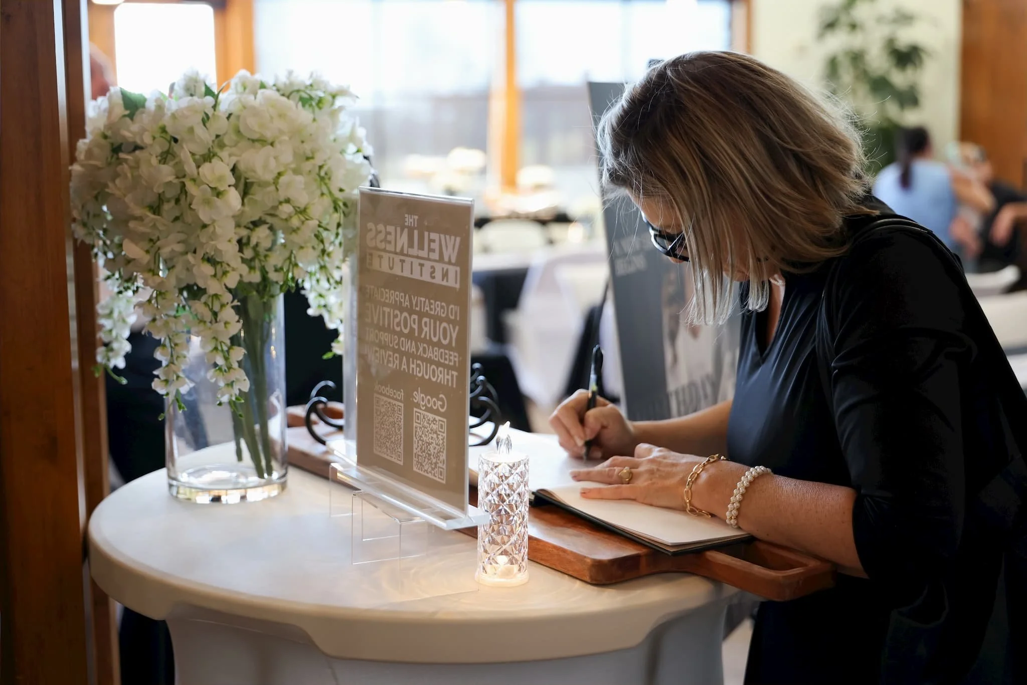 A woman wearing glasses and a black dress signing a guest book at a table with a large white floral arrangement and a sign with a QR code on it.