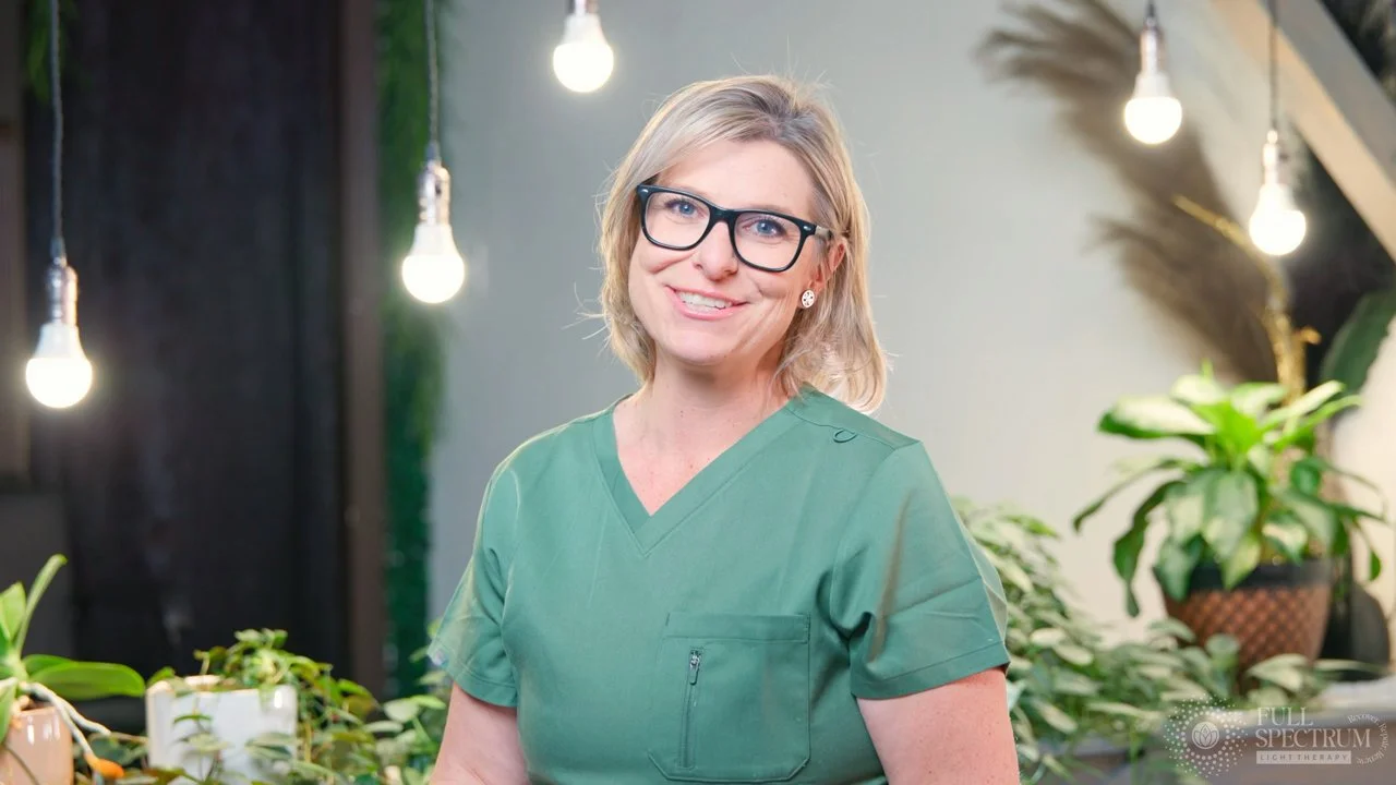 A smiling woman with short blonde hair, glasses, and a green medical uniform standing in a well-lit room with hanging light bulbs and potted plants.
