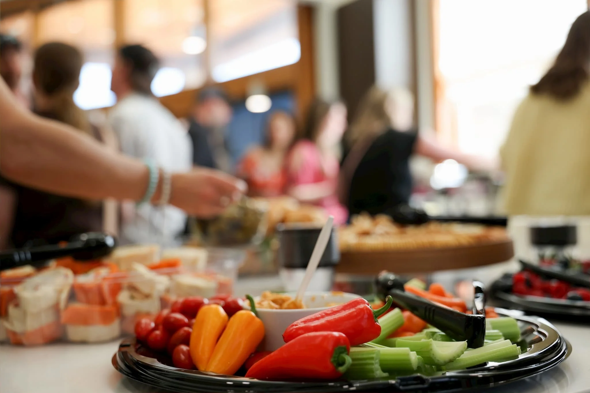 Colorful vegetables including red peppers, celery, cherry tomatoes, and baby carrots on a buffet table, with blurred people in the background.