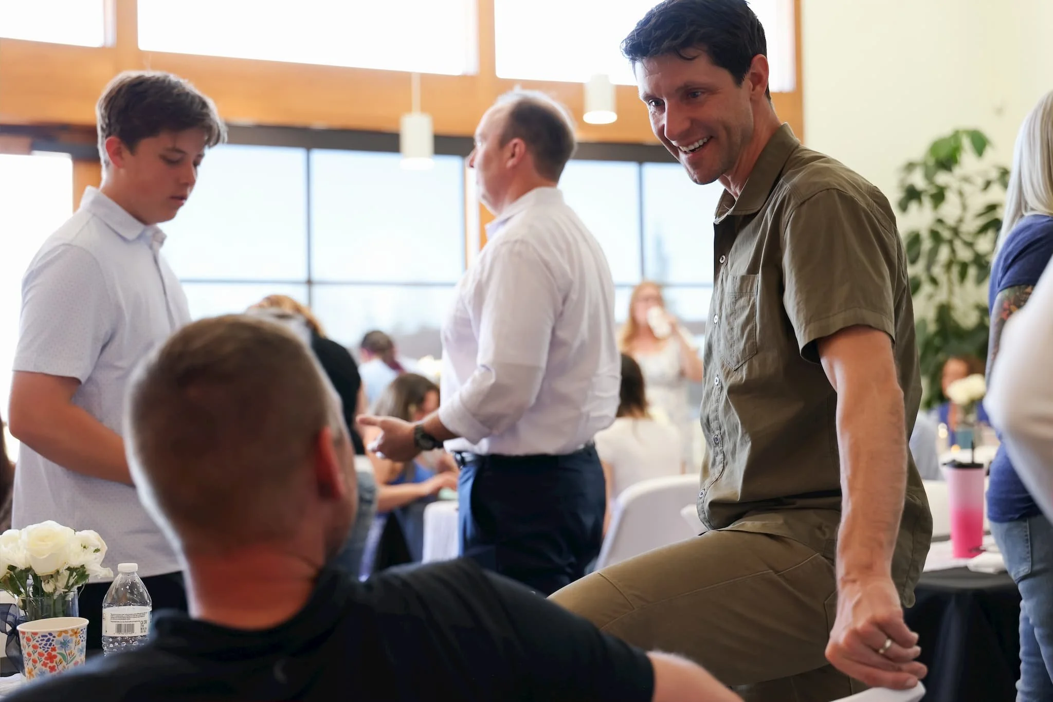 A smiling man in a beige shirt holding hands with others in a social gathering or event in a bright room with large windows.