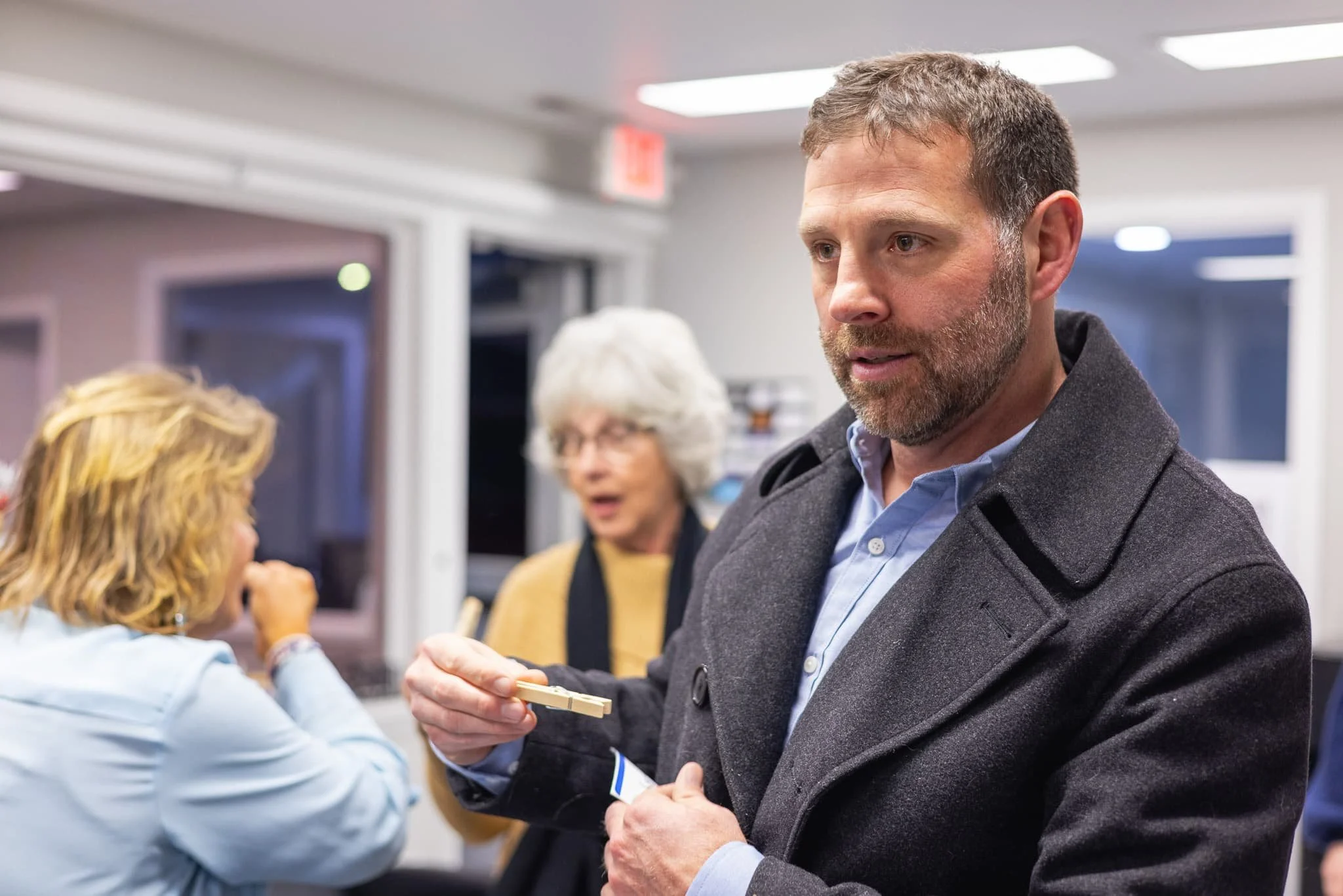 A man with a beard and short hair wearing a dark coat and light blue shirt interacts with a woman with blonde hair, who is wearing a light blue shirt, in an indoor setting that appears to be a public or office space. Two other women are blurred in th