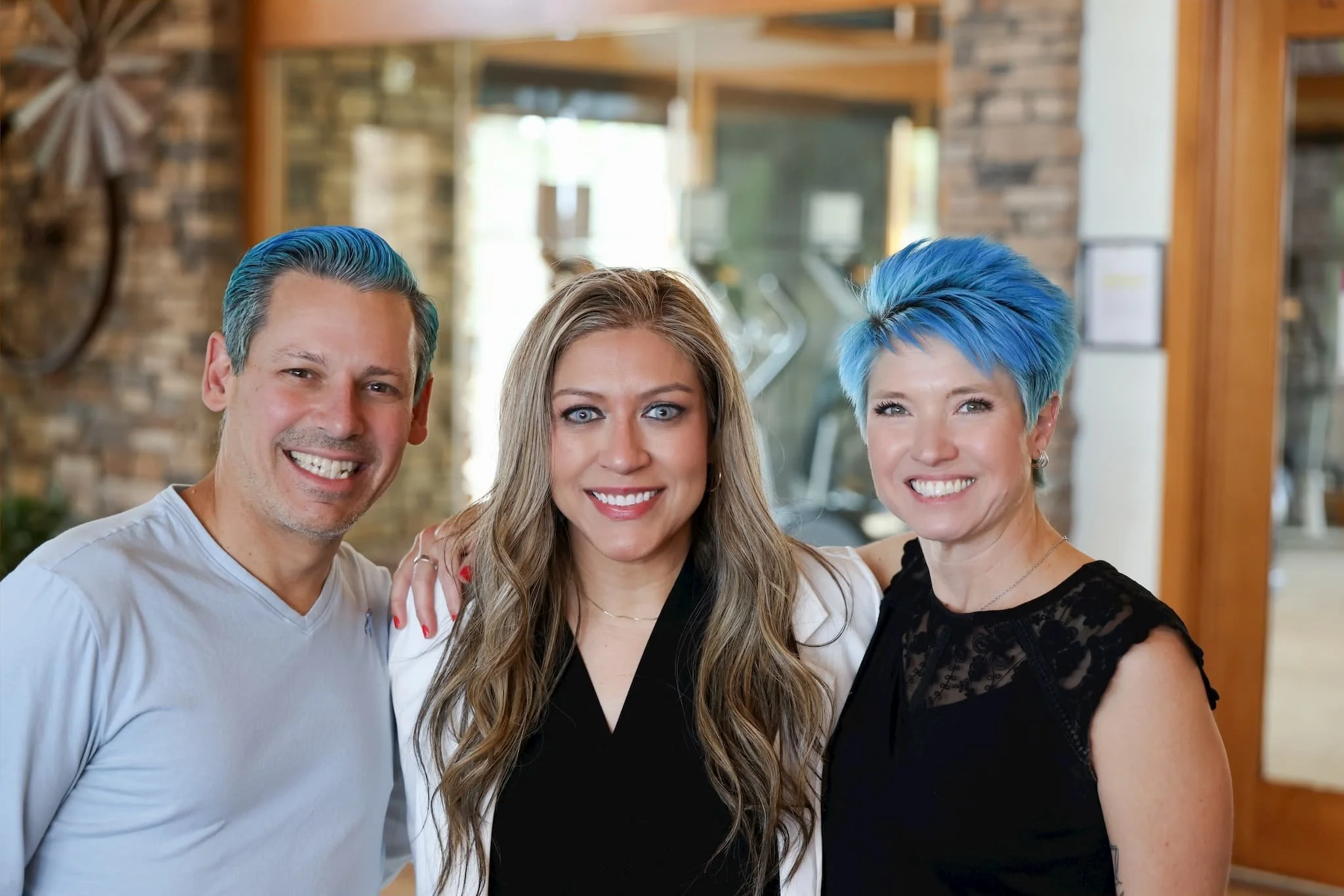 Three people with blue hair smiling inside a house or restaurant, standing close together, with brick and wood decor in the background.