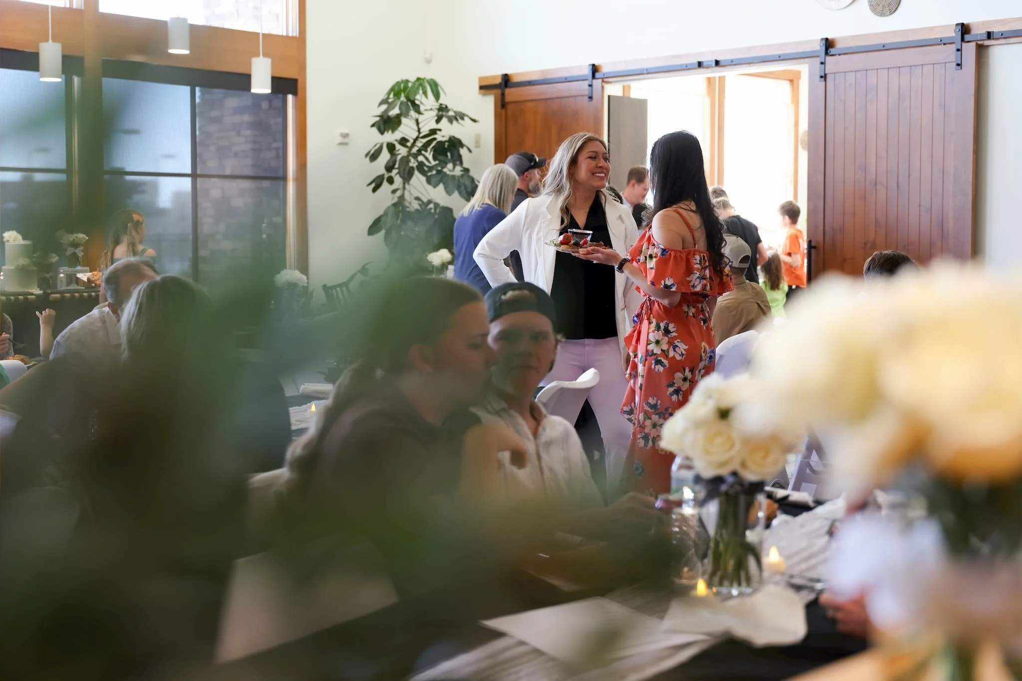 People gathered at a social event, with two women in the center smiling and talking, one holding a tray with food. The setting appears to be indoors with wooden sliding barn doors and contemporary decor.