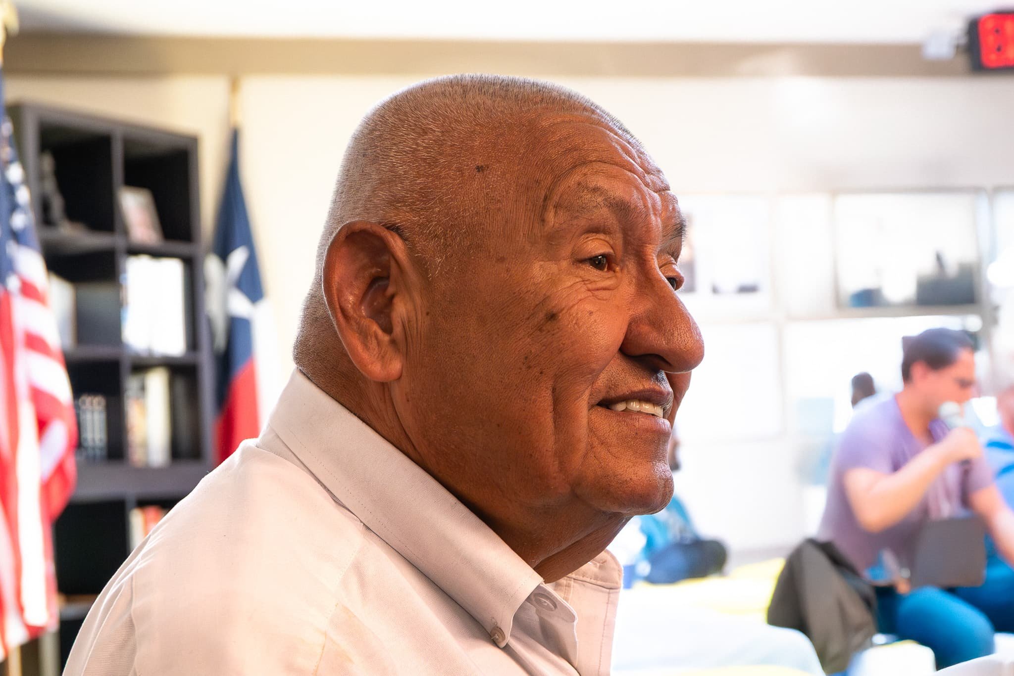 Close-up side profile of an elderly man with short gray hair and a dark skin tone, smiling while wearing a white shirt in a well-lit indoor setting with people and American flags in the background.