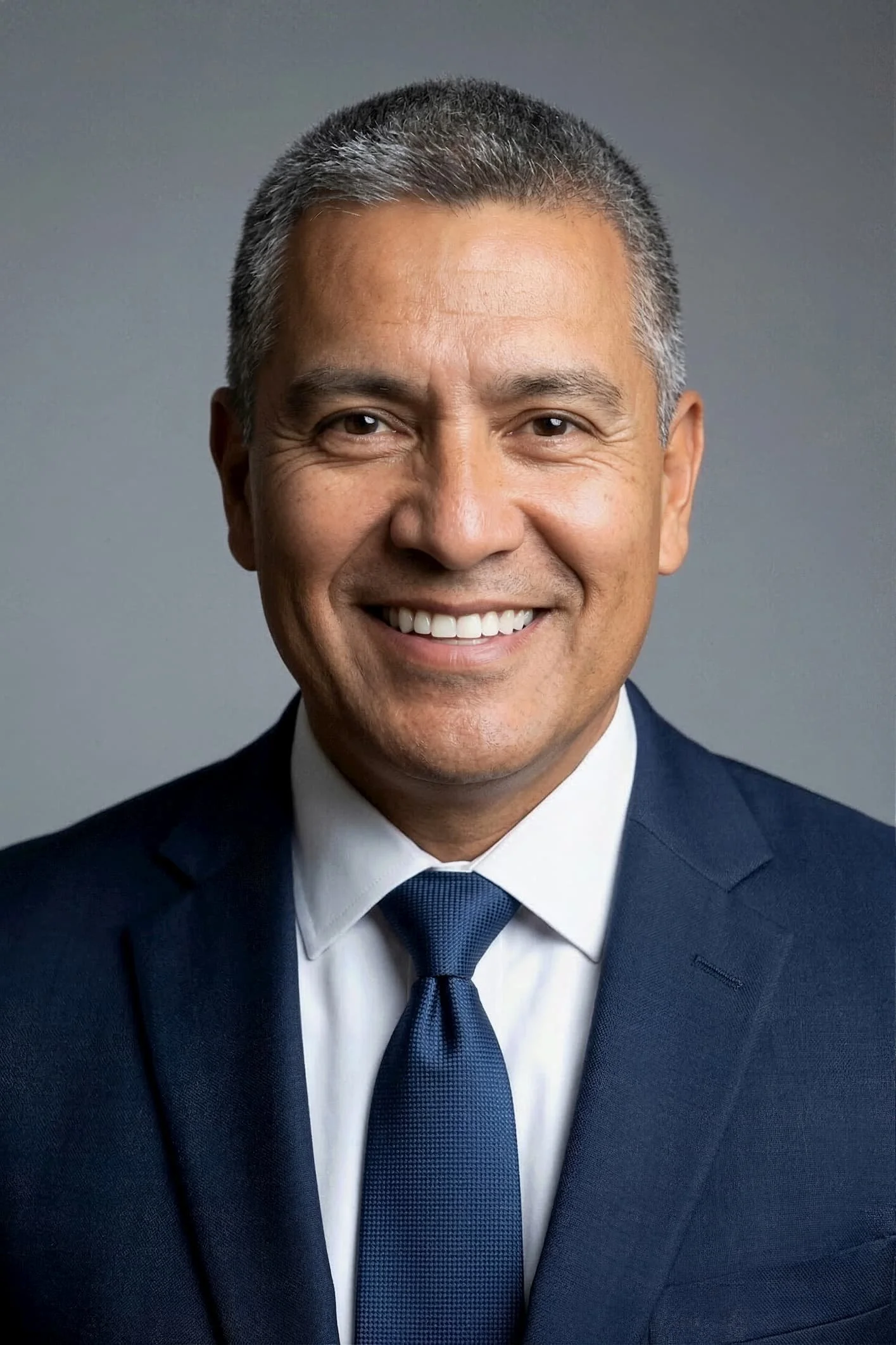 Close-up of a smiling man in a dark blue suit, white shirt, and blue tie, against a gray background.
