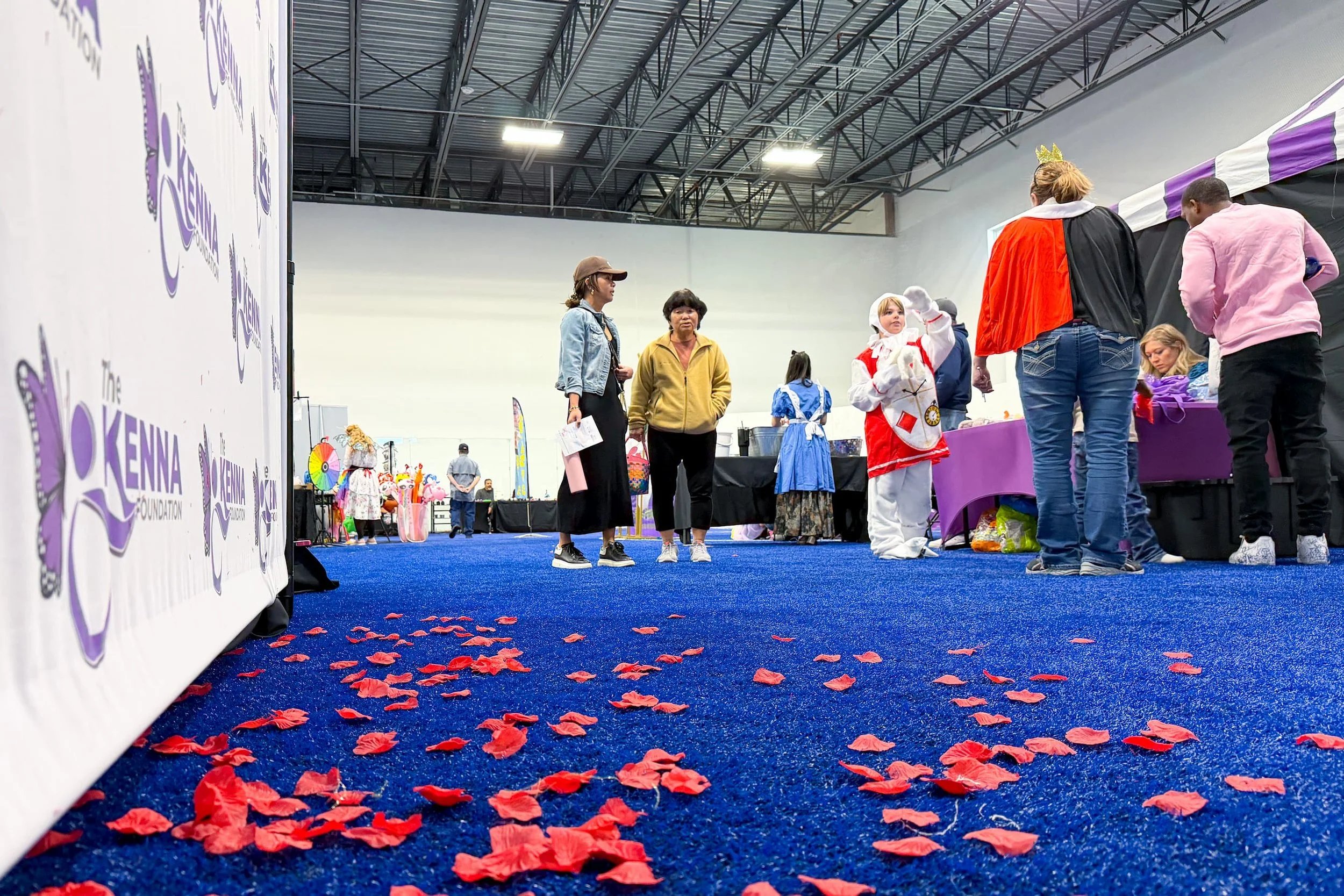 People at an indoor event with a blue carpeted floor decorated with red flower petals, and a large white banner with purple logos on the left side. Several individuals are standing in front of a purple table, including a girl in a white and red bunny