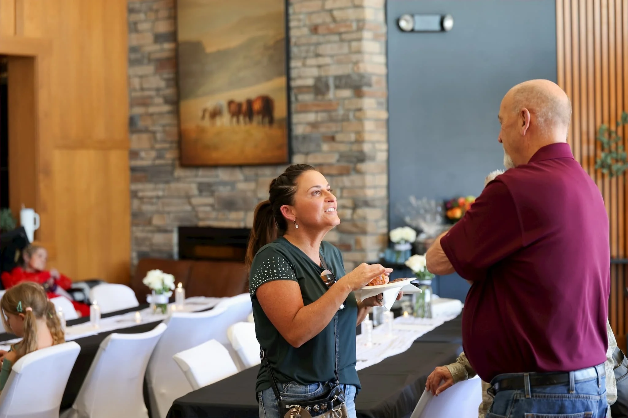 A woman with dark hair in a ponytail wearing a dark green shirt with rhinestones is serving food to a bald man with a gray beard wearing a maroon shirt at a social gathering indoors. There are white chairs and tables with white tablecloths in the bac
